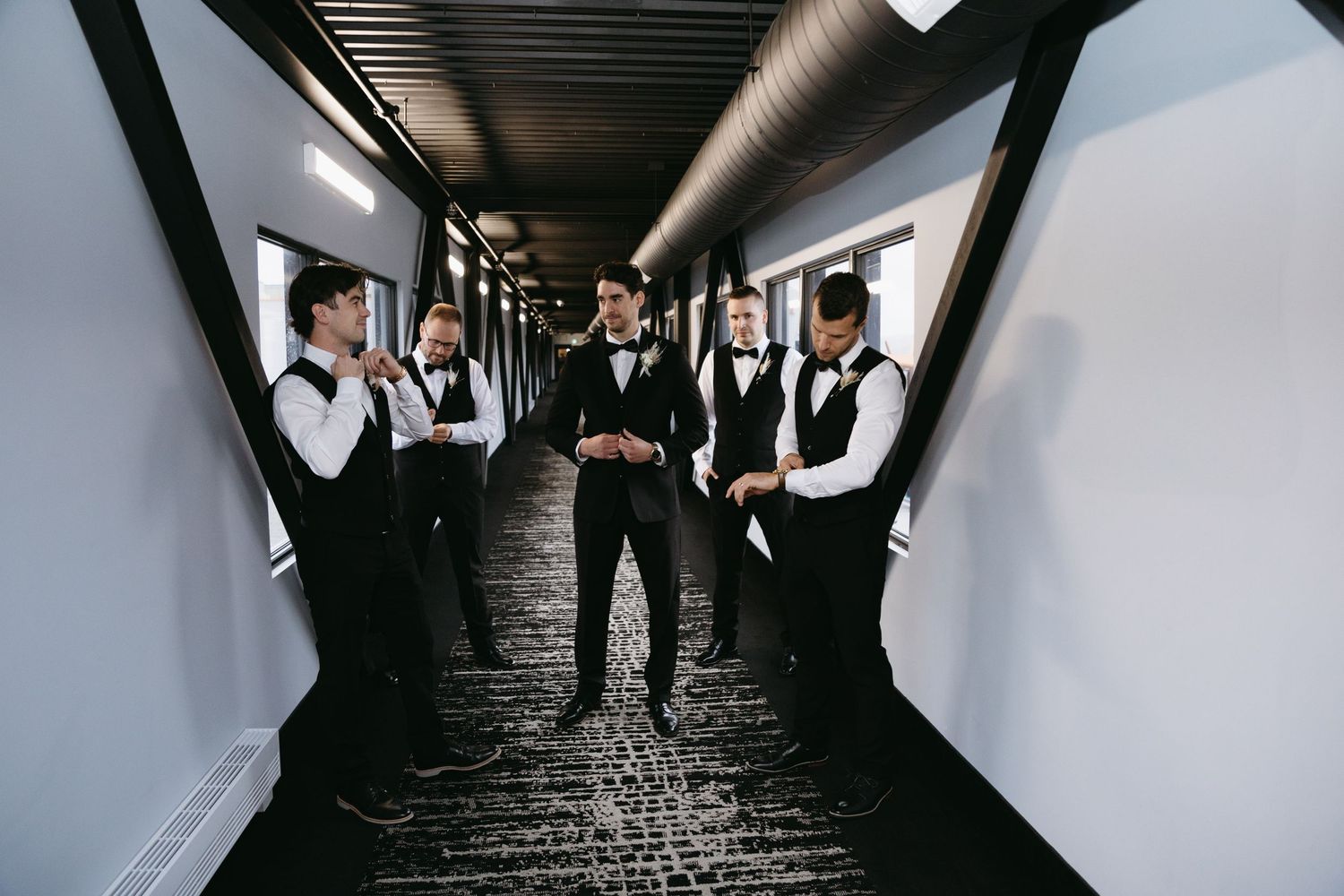 A group in formal black and white attire stands in a dramatic corridor with industrial lighting overhead.