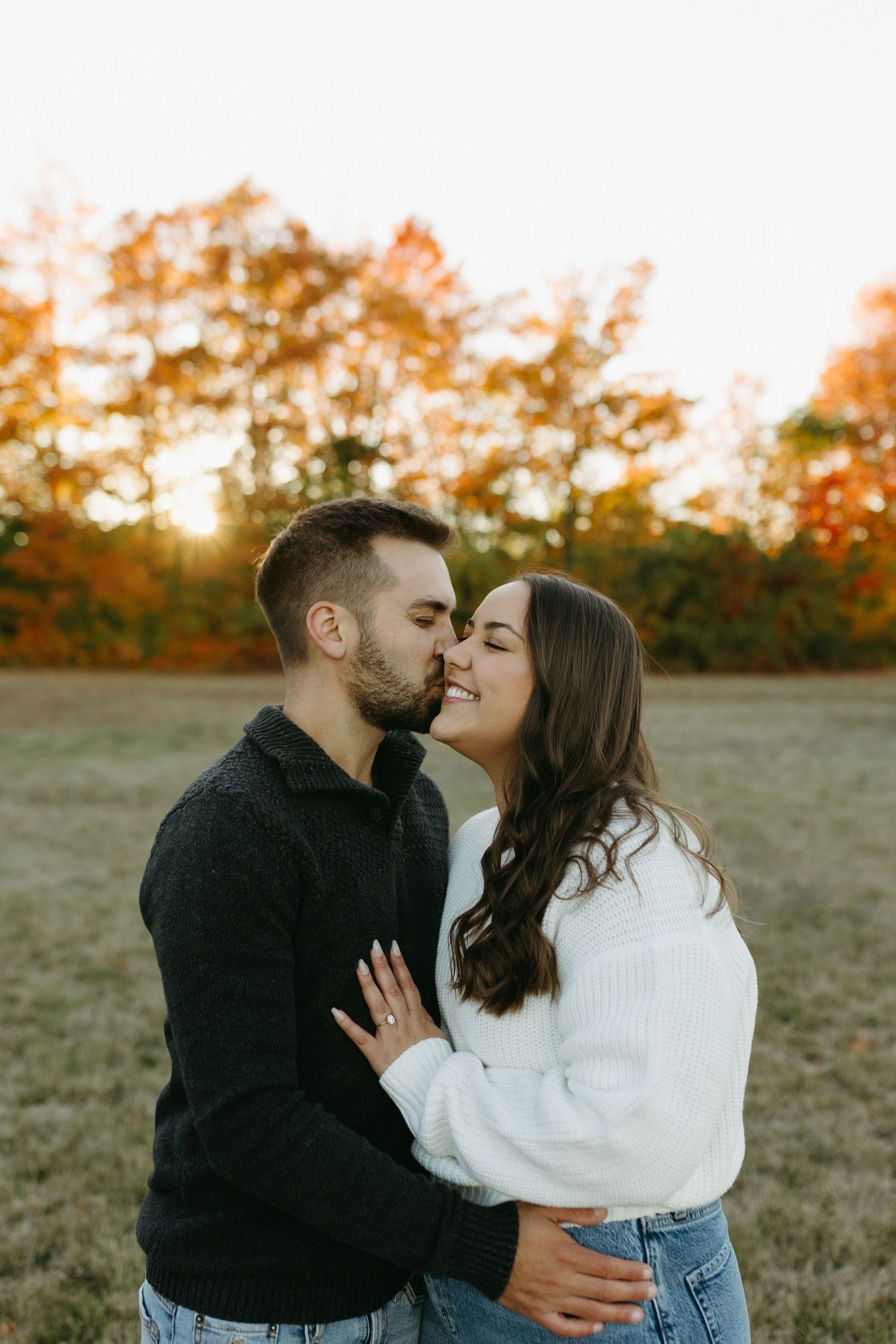 A romantic couple shares tender moments in a field during golden hour with autumn trees in the background.
