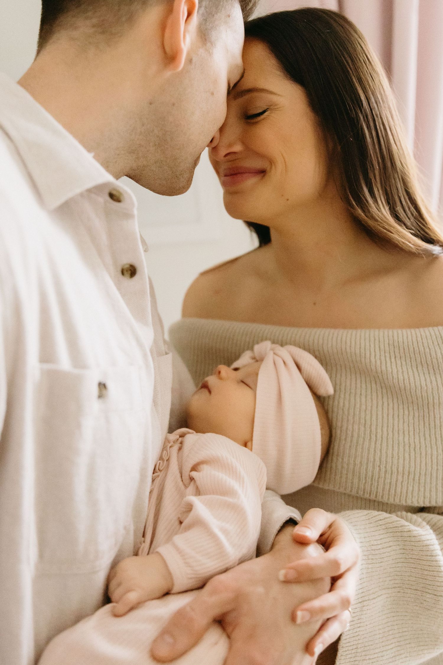 A tender moment between parents holding their newborn baby in soft natural light against a light backdrop.