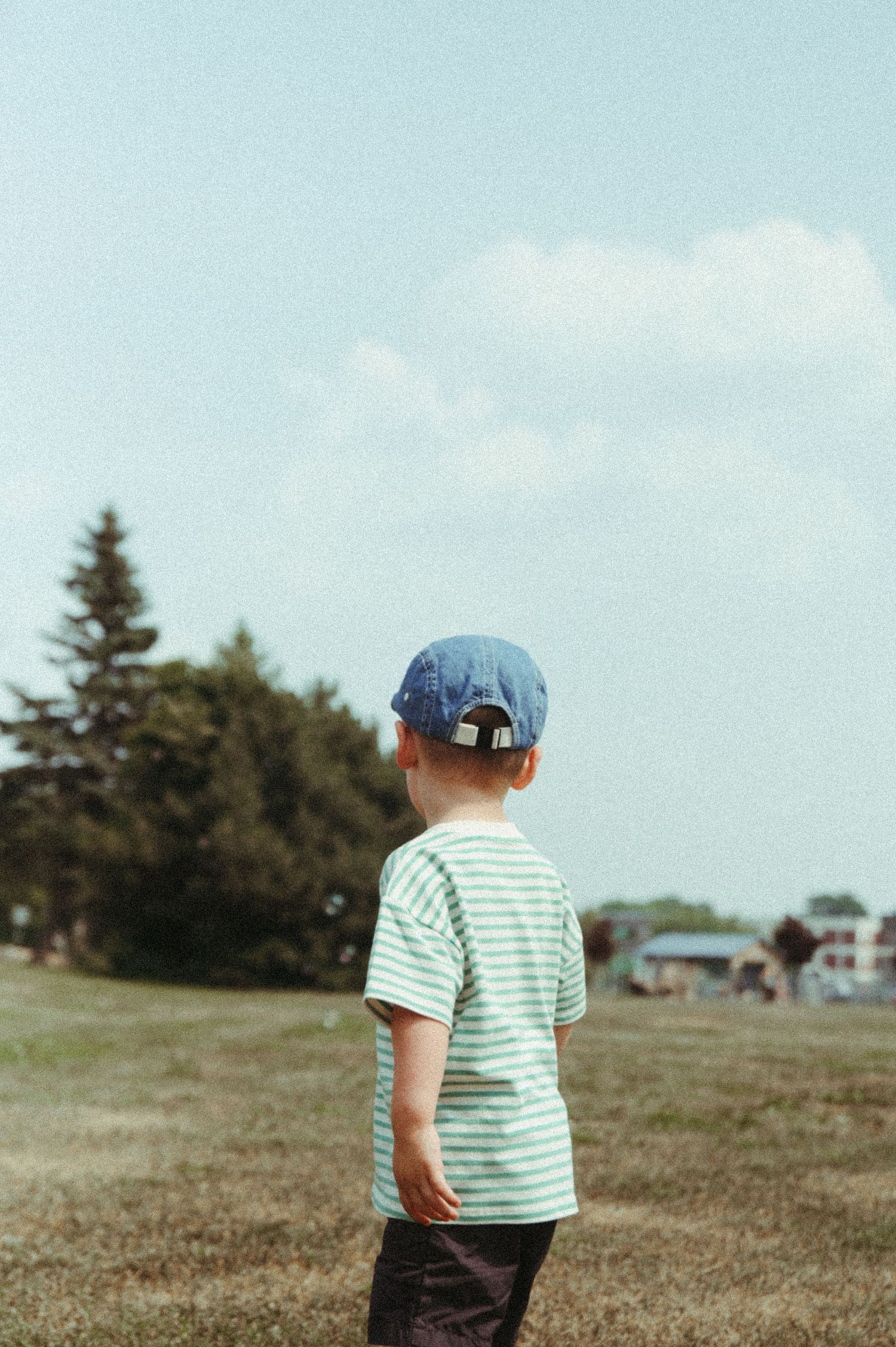 A small child in a striped shirt and blue hat standing in a grassy field under clear blue skies.