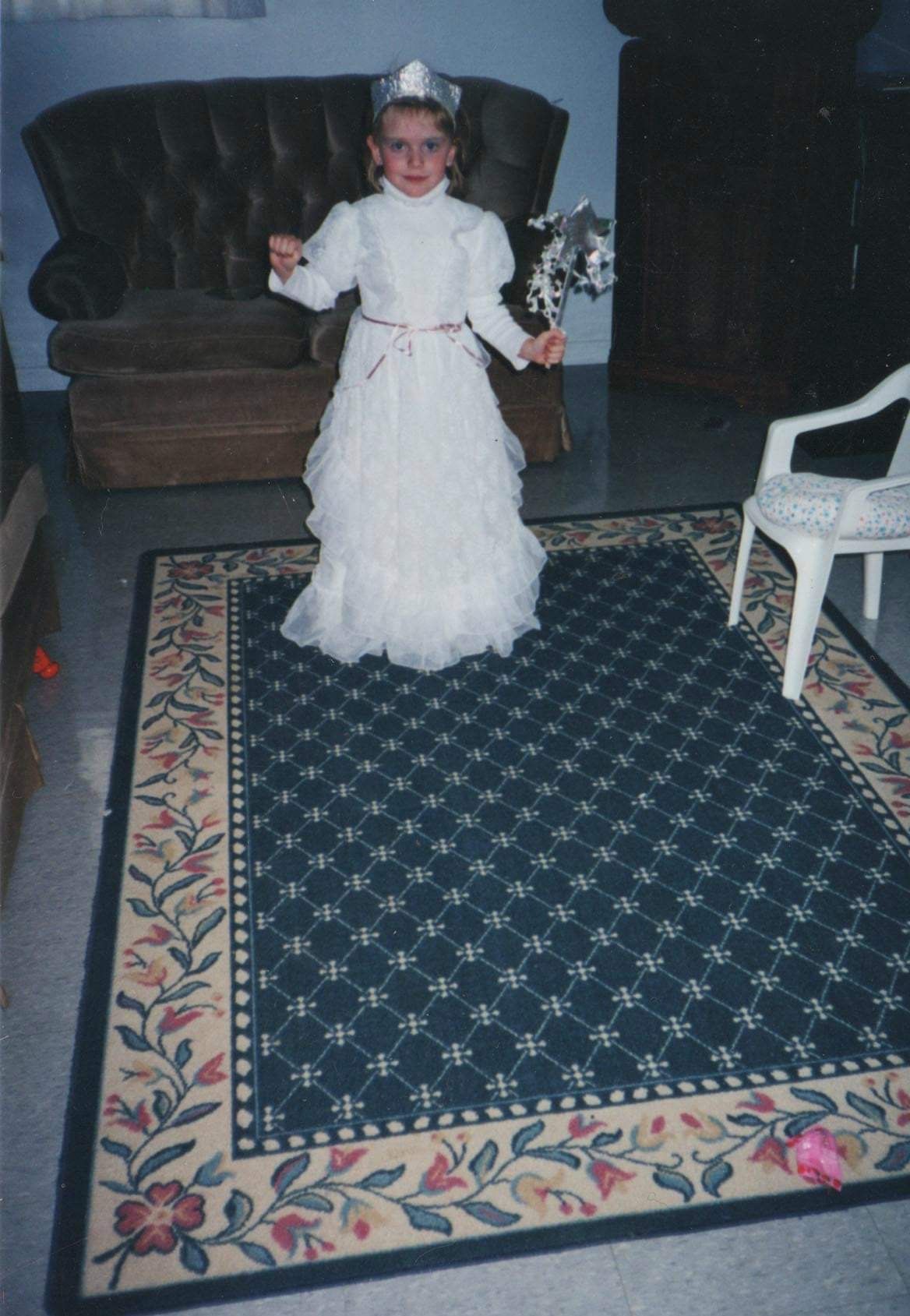 A small child in a white dress stands on a dark blue area rug with floral border in what appears to be a living room.