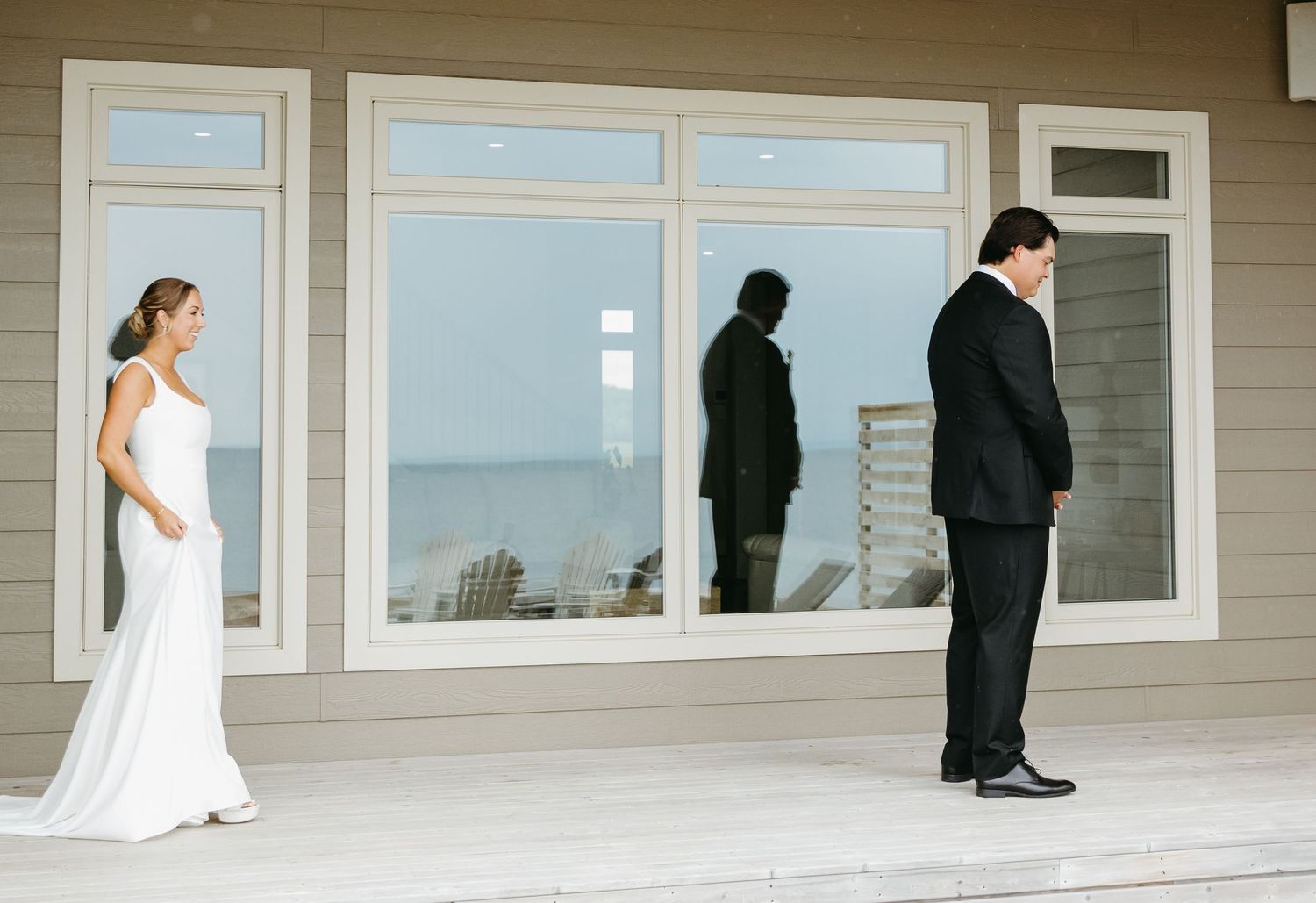 A creative panoramic wedding photo shows a couple walking apart on a covered porch with large windows.