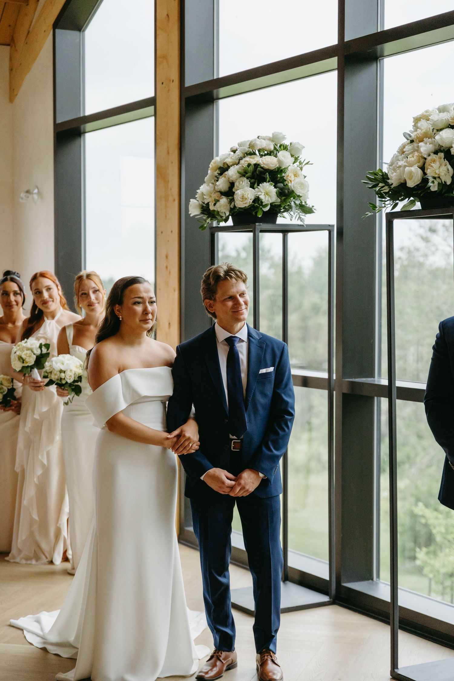Wedding party members in white dresses and navy suits celebrate joyfully by large windows in modern venue.