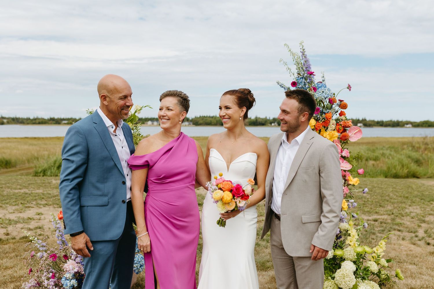 Wedding party laughs together on a scenic beach with bright flowers and coastal grasses in the background.