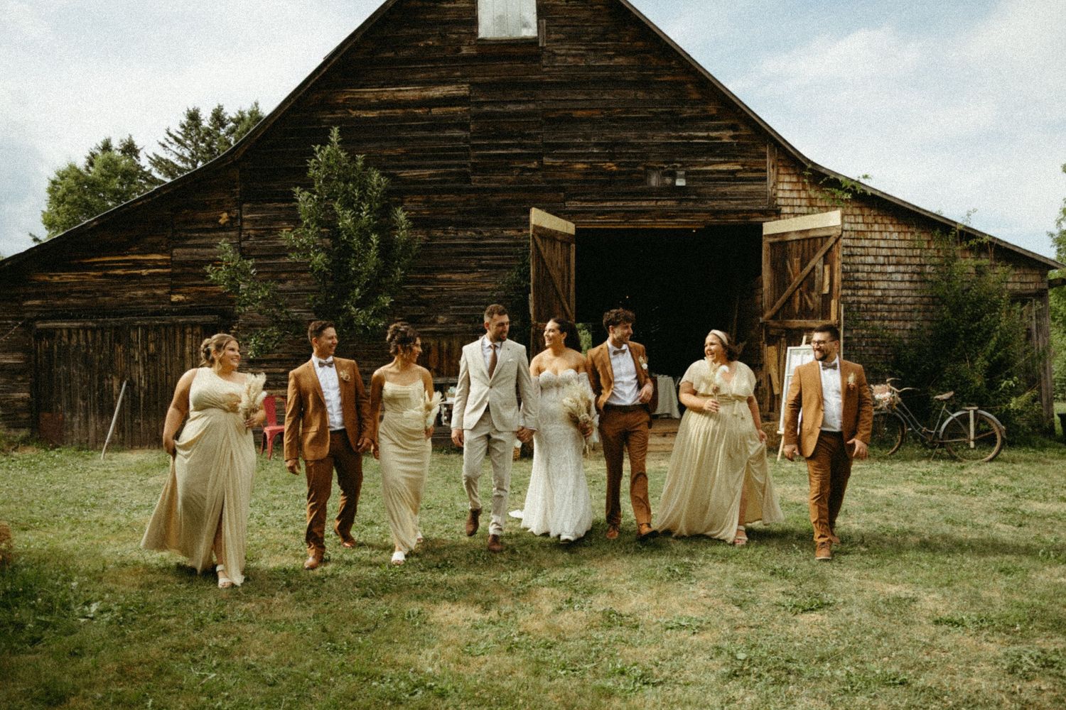 Wedding party walks together in a line across rustic farmland with weathered wooden barns in the background.