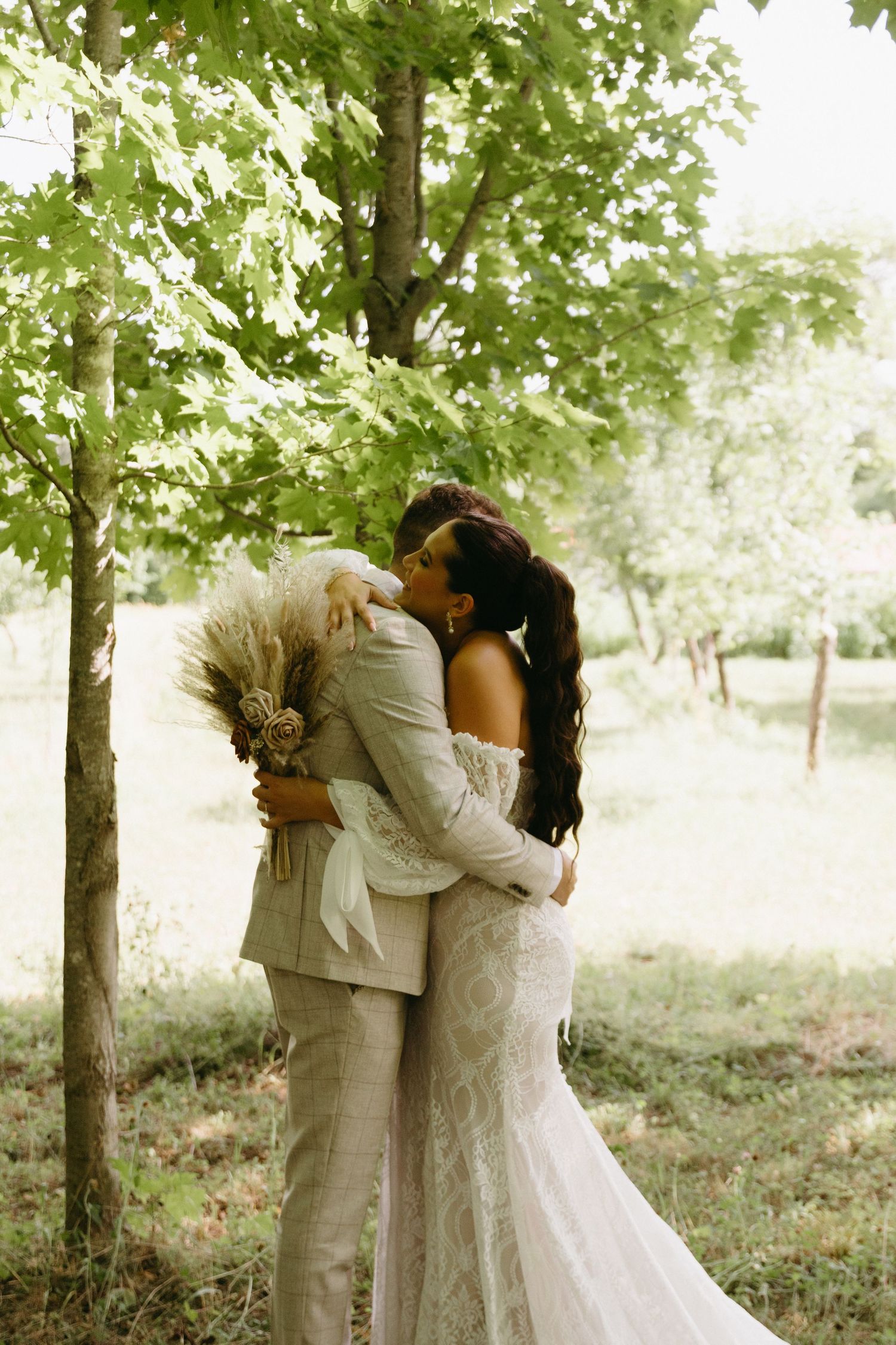 A romantic wedding couple shares an intimate kiss in a serene wooded setting with sunlit trees in the background.