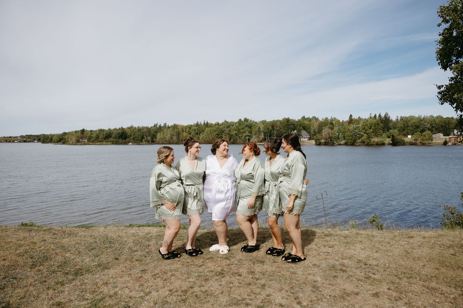Group in white outfits poses playfully on a beach with lake and trees in the background.