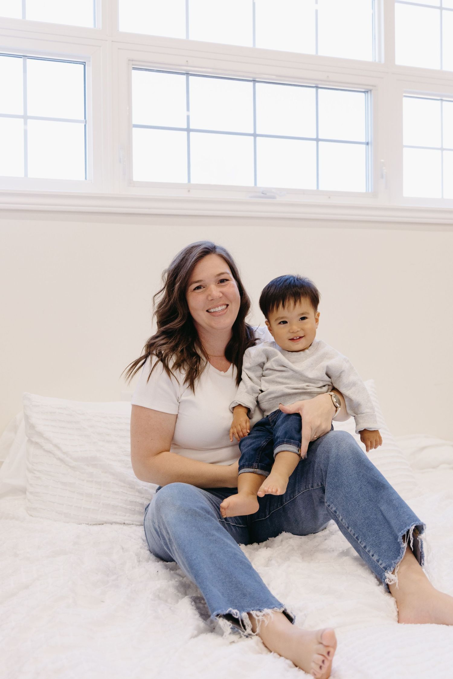 A parent and child sit together on a white carpet in a bright sunlit room with large windows.