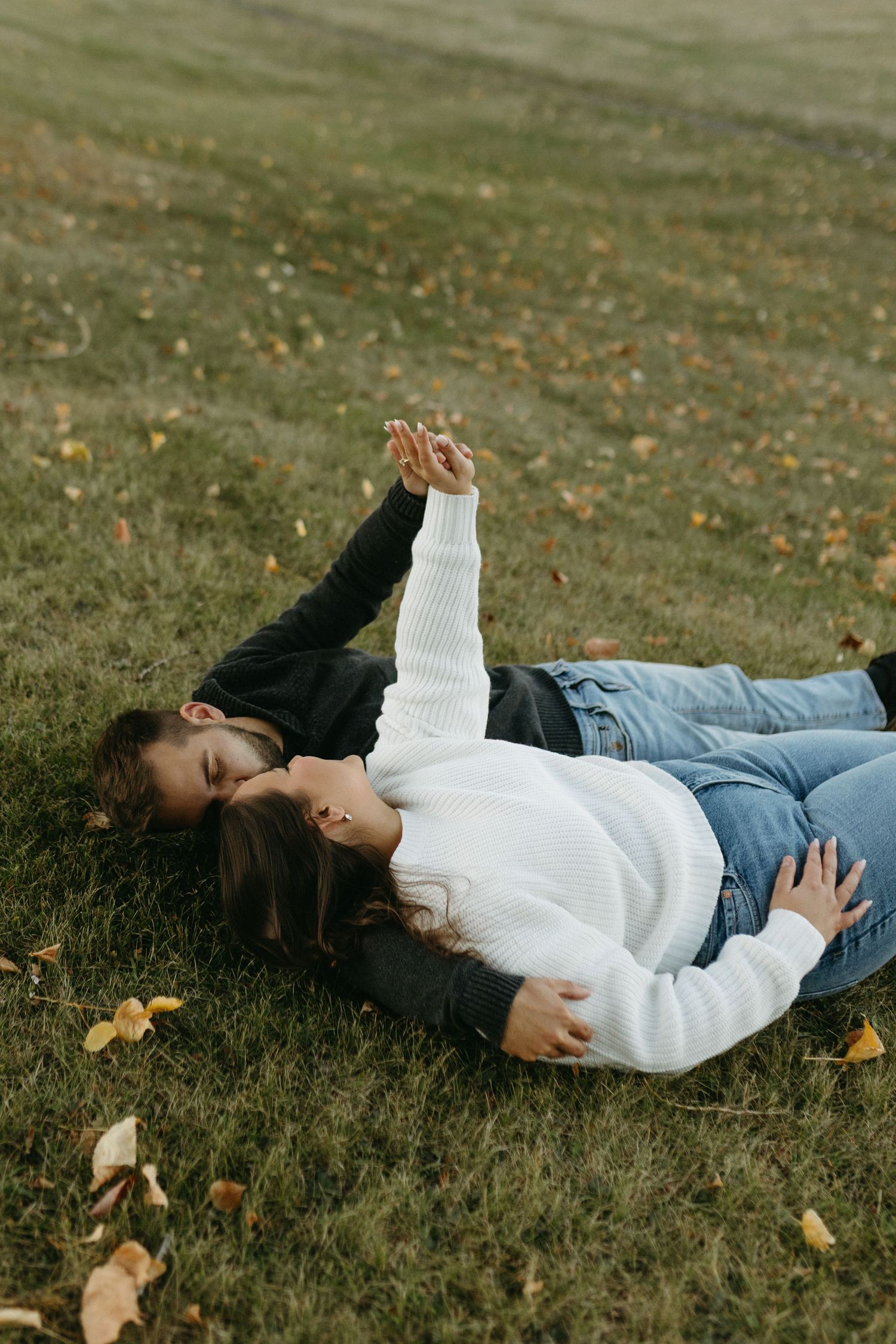 Person lying on grass wearing white sweater and jeans surrounded by scattered autumn leaves in a peaceful outdoor setting.