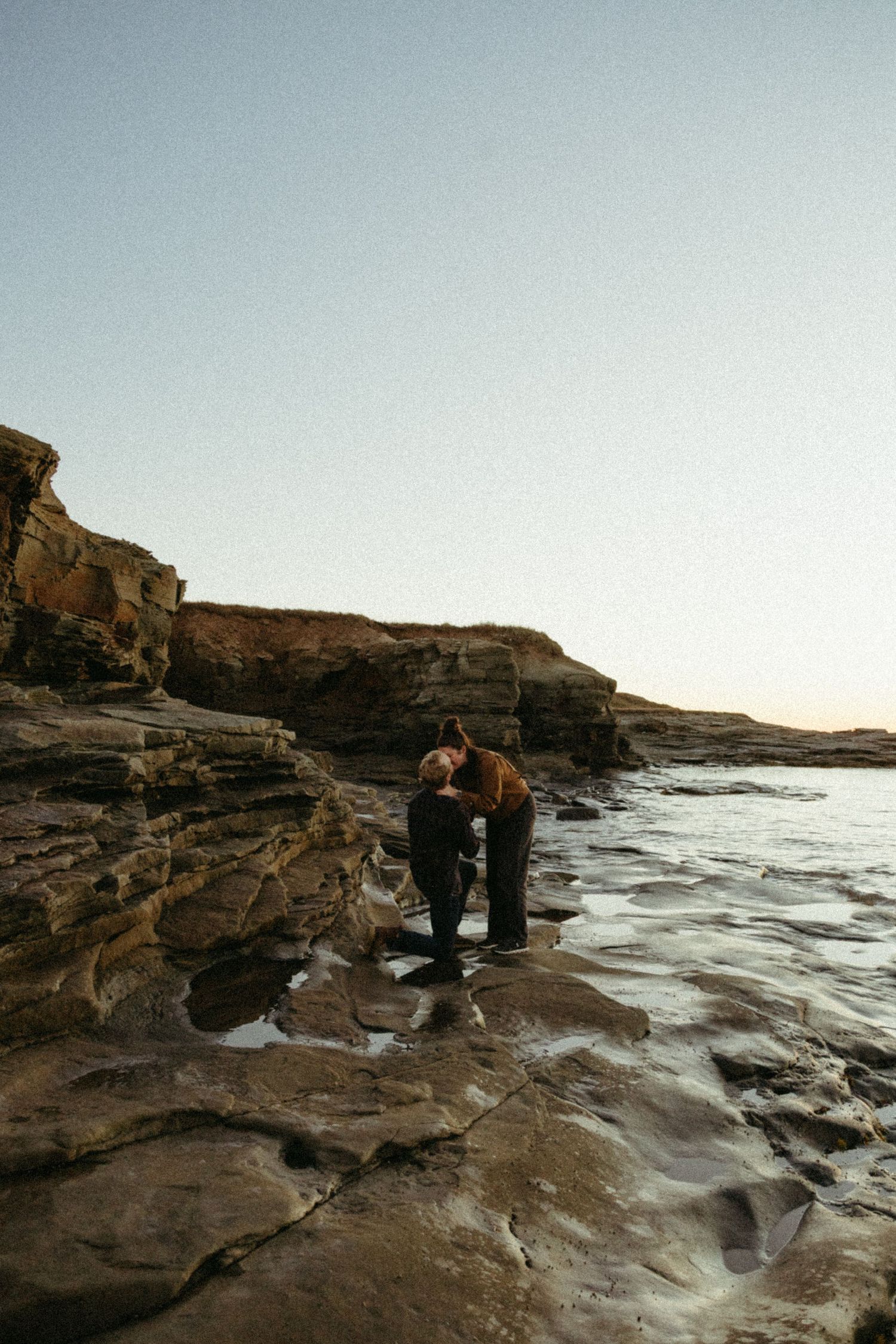 Two people share a tender moment on rocky coastal cliffs at sunset as waves crash against the shore.