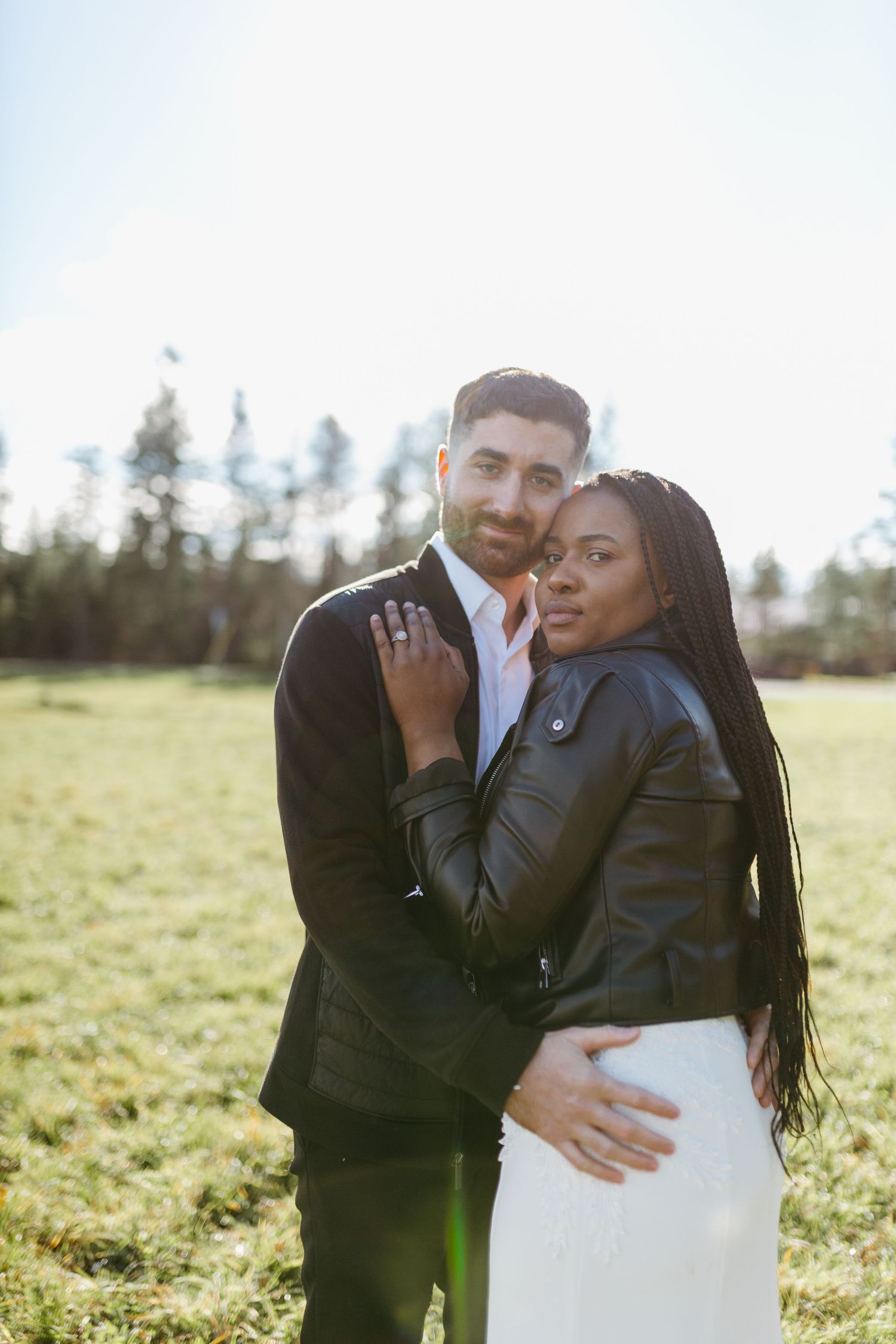 A couple poses together in a sunny field, with her wearing a leather jacket and white dress.