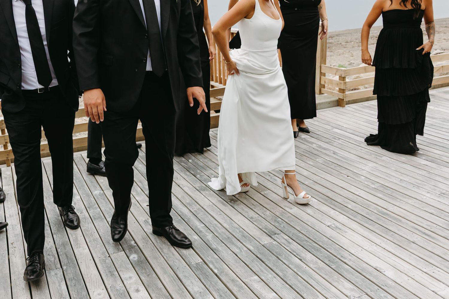 Wedding party members in formal attire stand together on a wooden deck during an outdoor celebration.