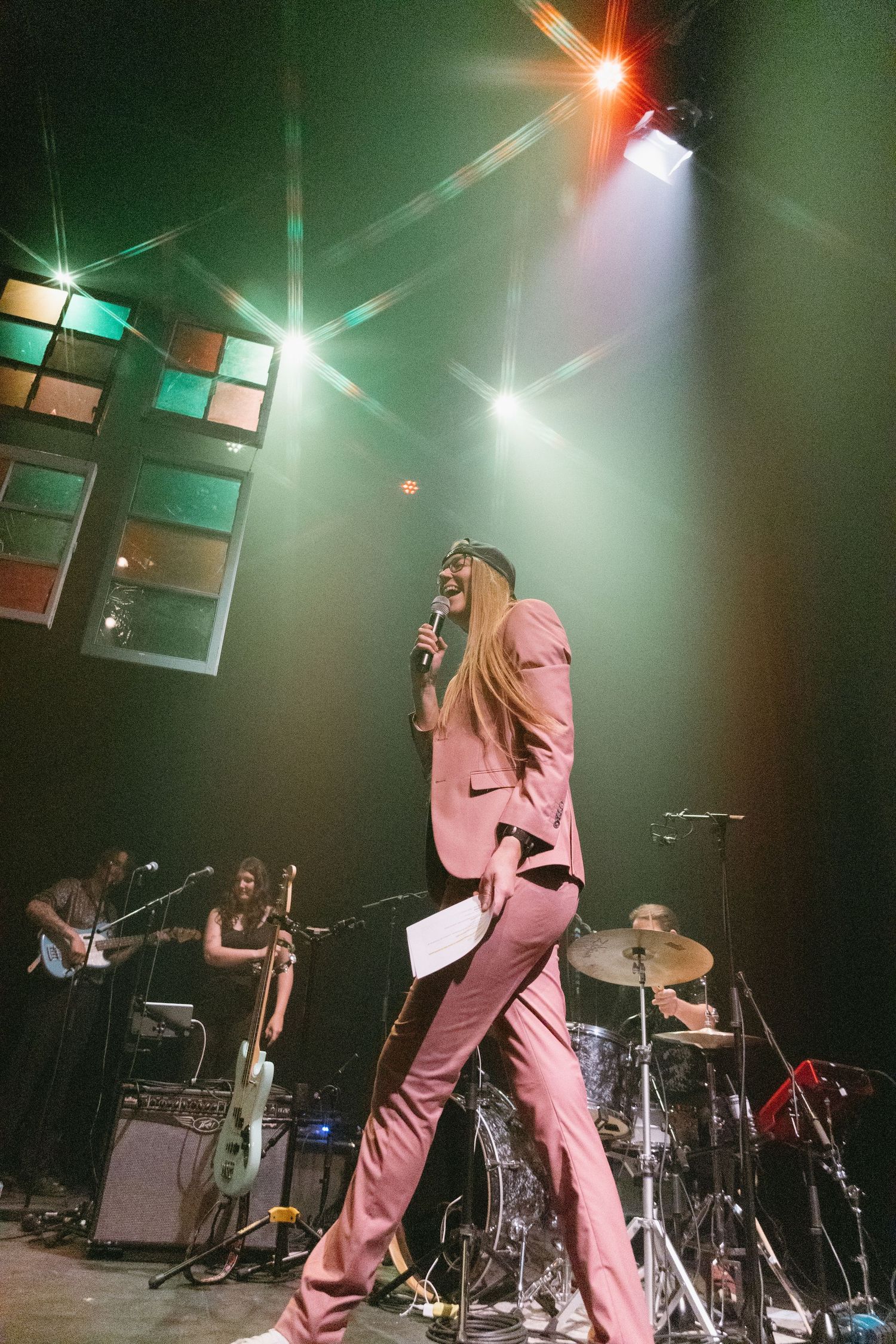 A performer in pink attire energetically moves across a dimly lit stage with dramatic lighting and musical equipment.