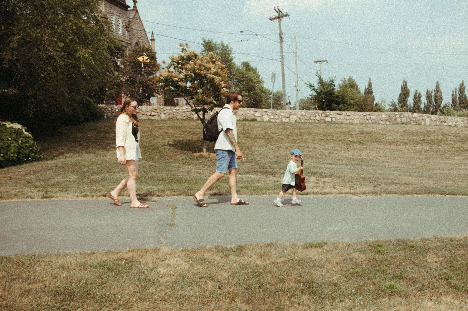 People walking along a grassy path on a sunny day with trees and utility poles in the background.