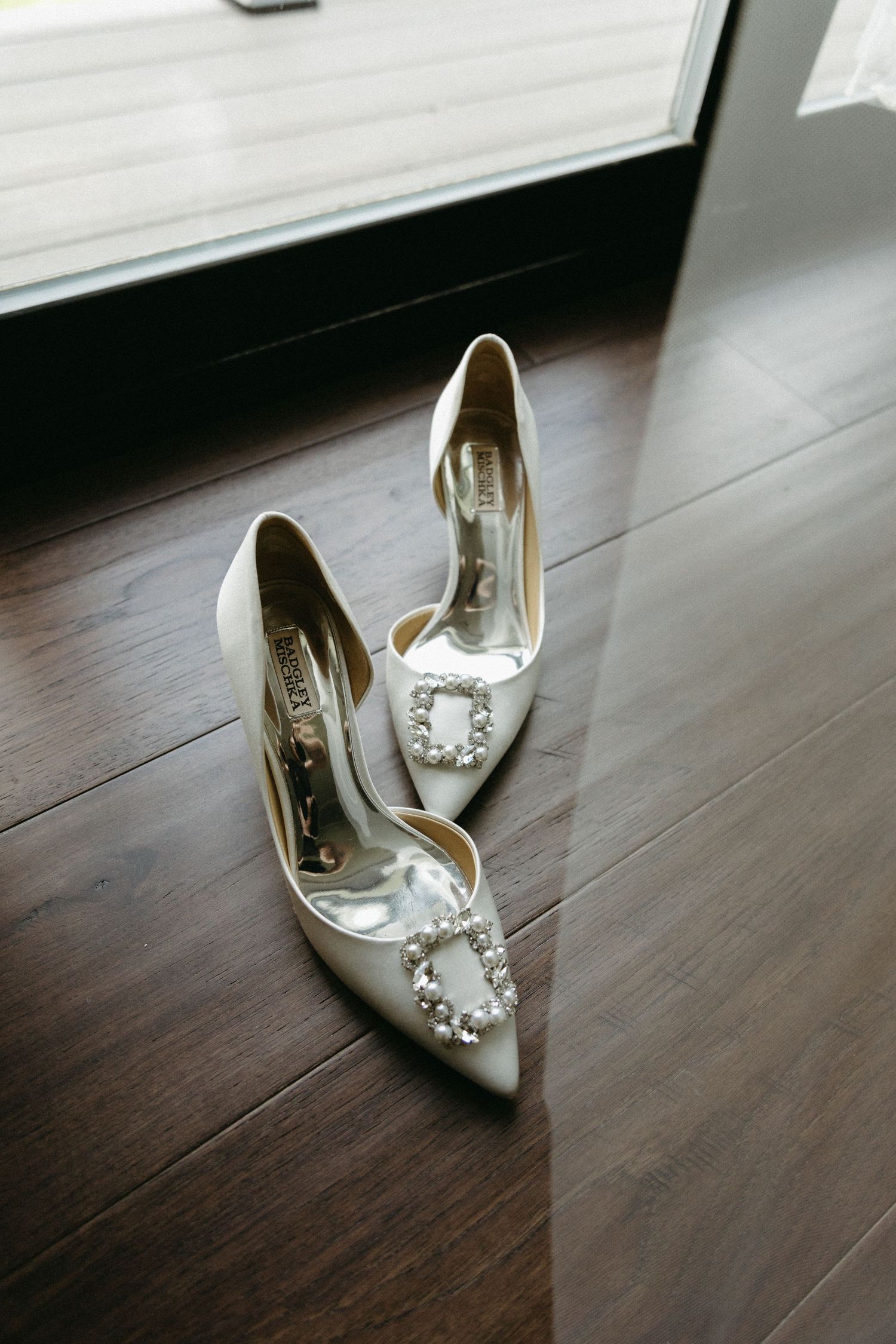 Elegant white satin bridal shoes with crystal embellishments sit on dark hardwood flooring by a window.