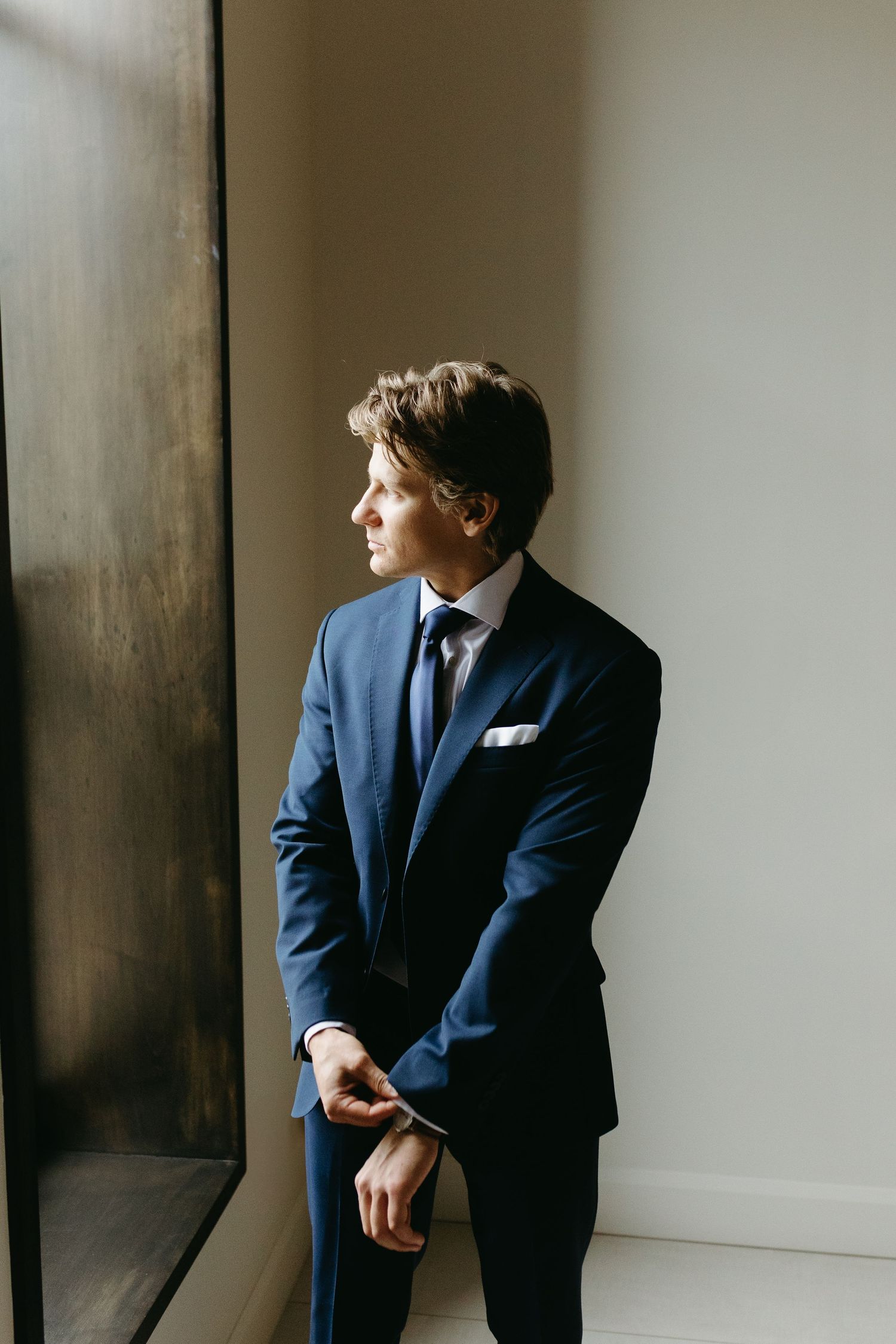 A person in a navy blue suit and tie stands by a window in contemplative pose, looking to the side in natural light.