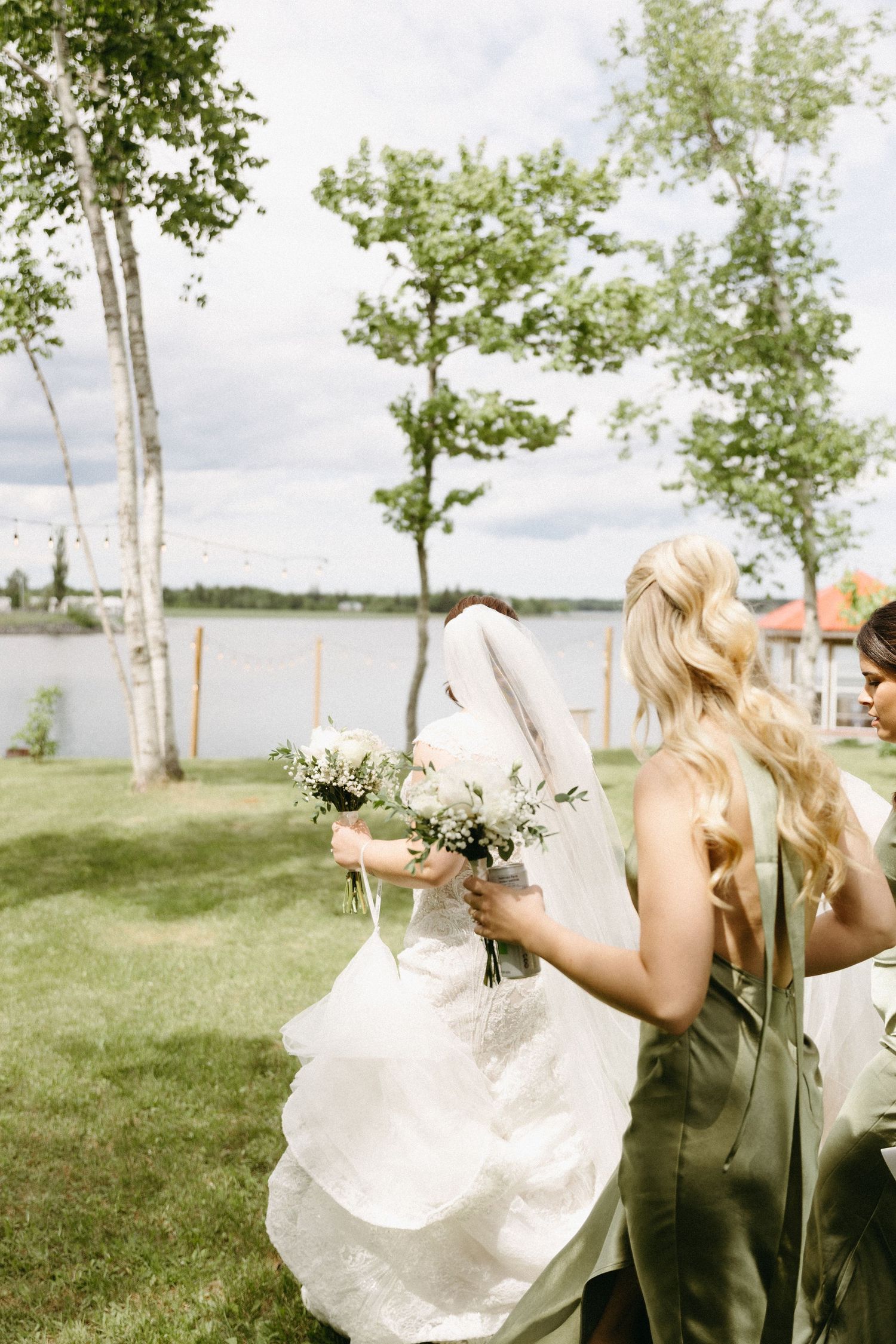 Two women in formal attire hold bouquets by a lakeside with birch trees and green grass on a cloudy summer day.