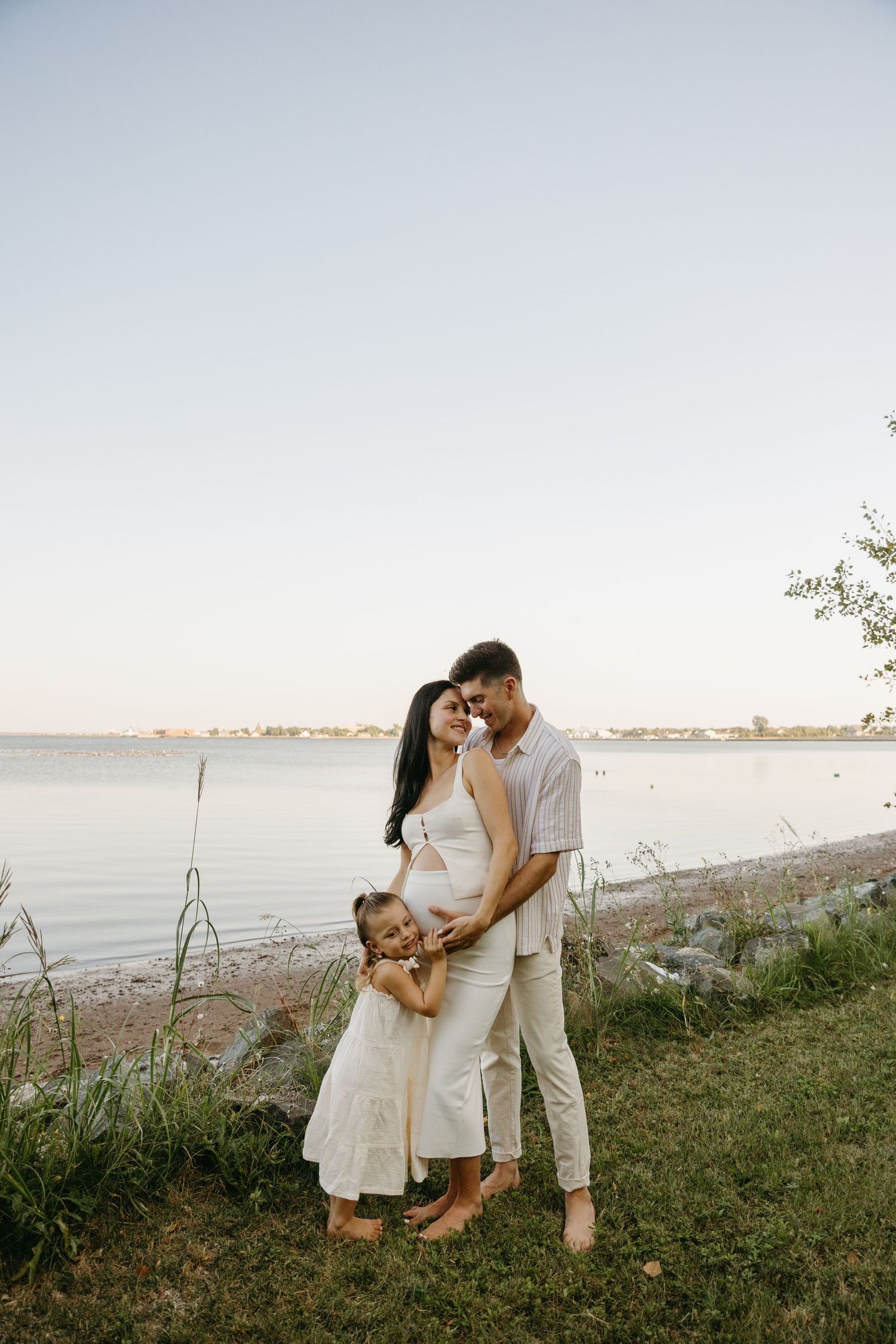 A romantic photoshoot at sunset showing a couple in white clothing embracing on a grassy hillside by the ocean.