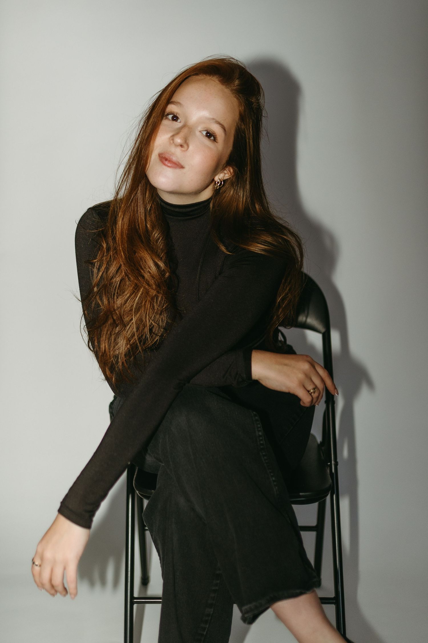 A person in a black turtleneck and dark pants poses on a chair against a white wall with dramatic shadows.