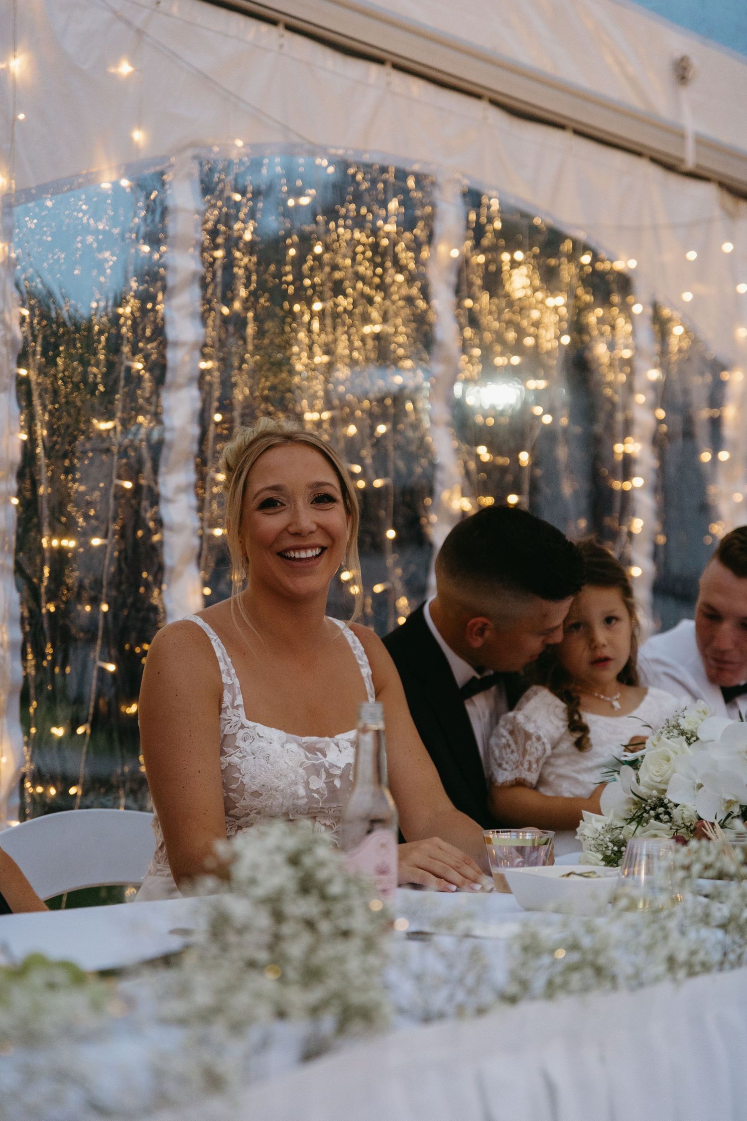 Wedding reception guests seated at tables under twinkling fairy lights in an elegant white tent setting.