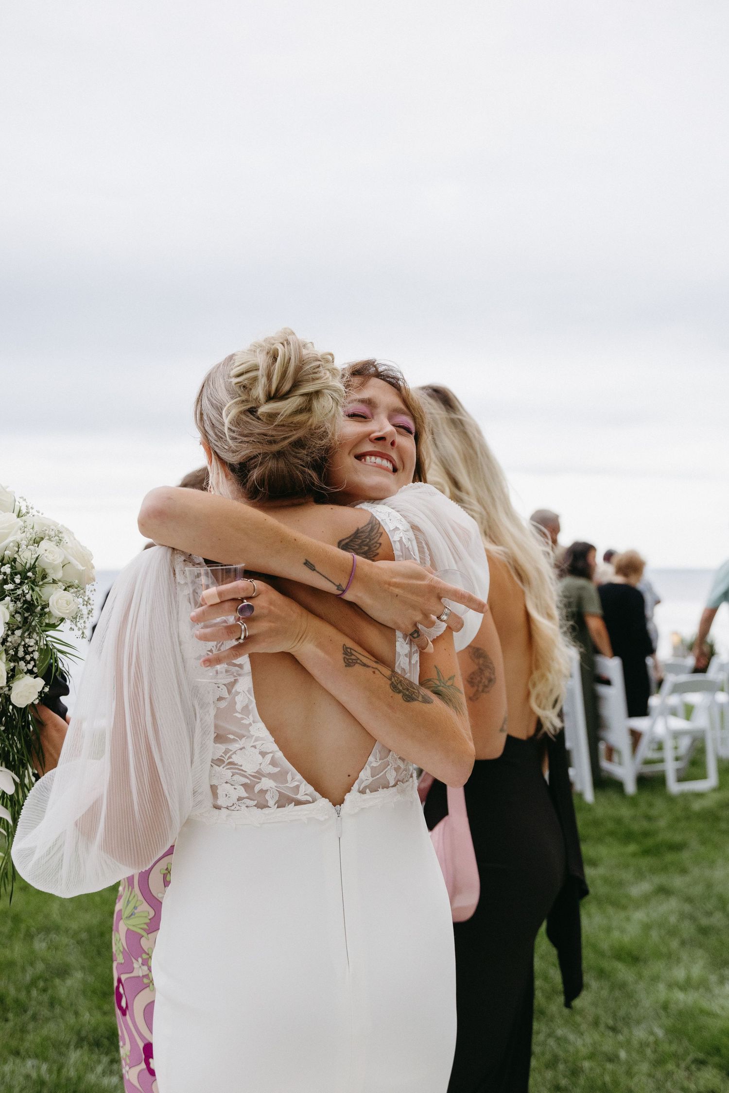 Two people share an emotional embrace during an outdoor wedding ceremony on a grassy lawn with white chairs in the background.