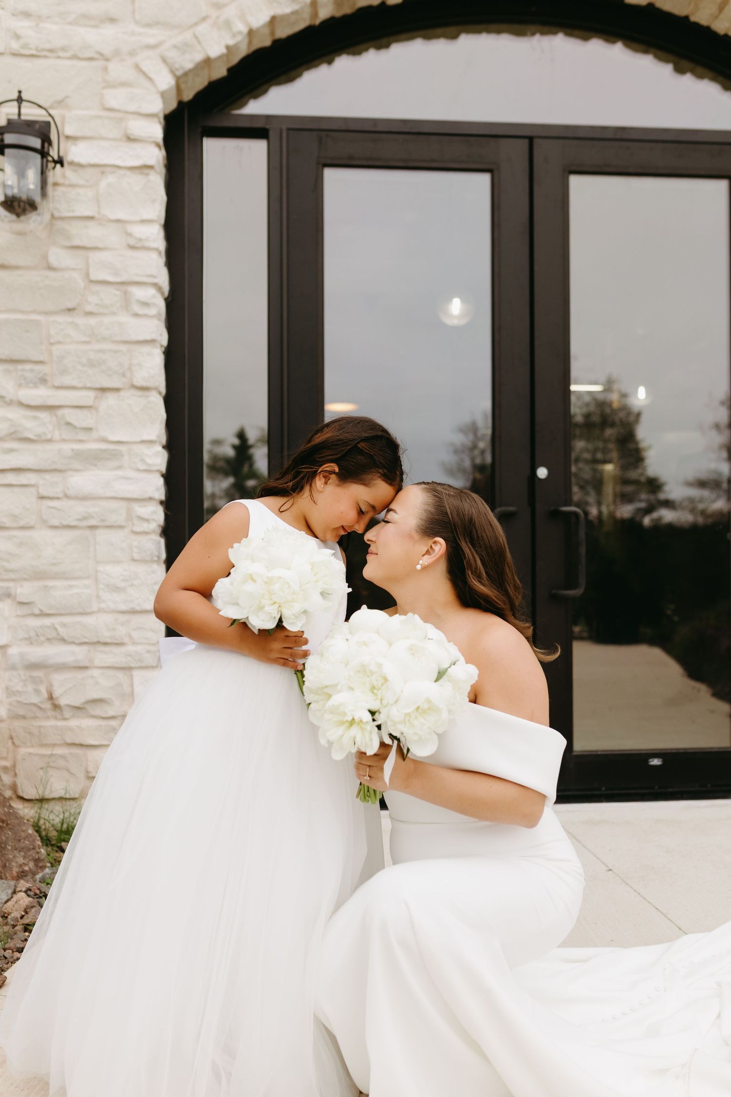 Two brides in elegant white wedding dresses share a romantic moment outside a modern stone building.