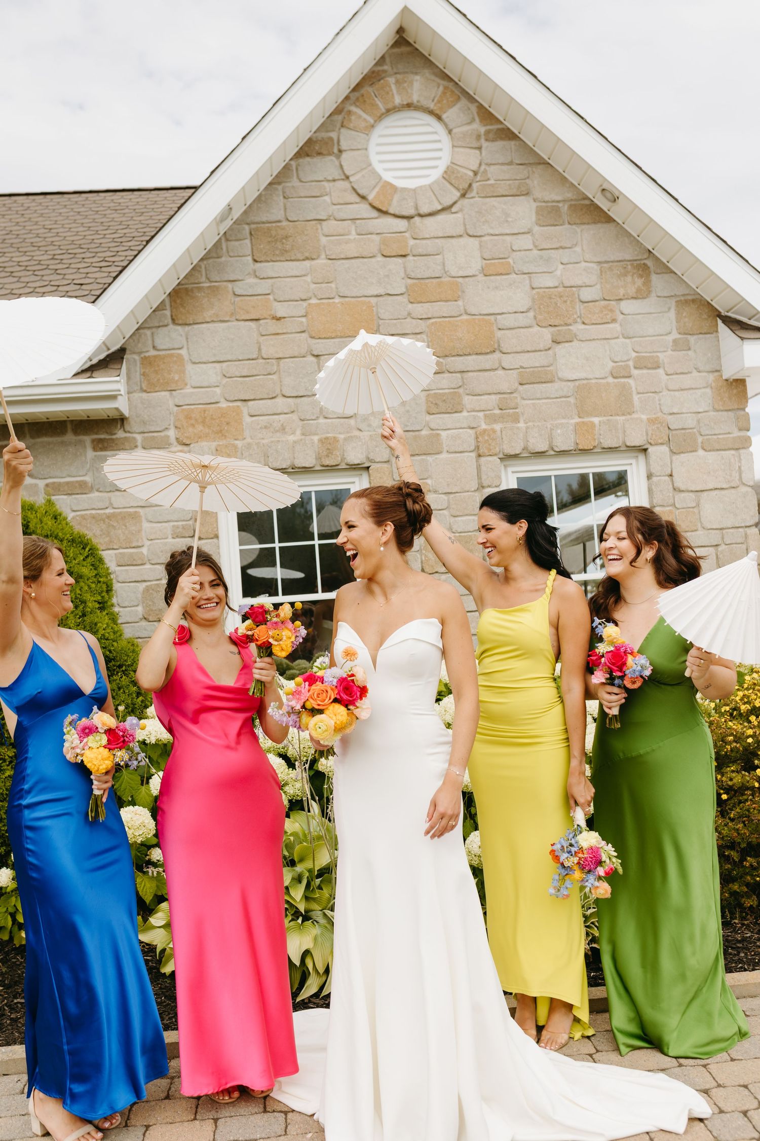 Bridesmaids in colorful dresses twirl and dance together in front of a stone building during a wedding celebration.