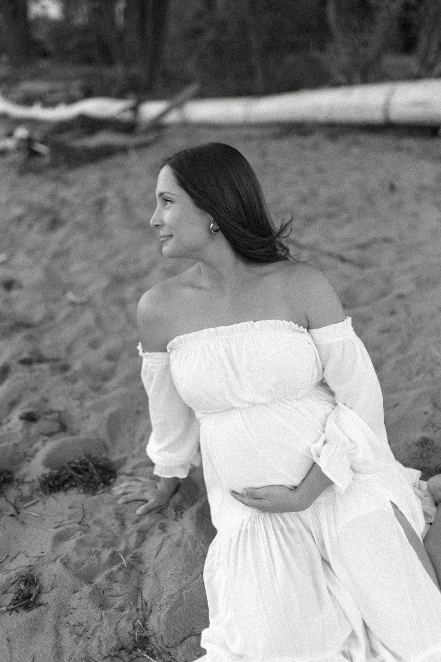 Woman in flowing white dress poses gracefully on a beach in black and white.