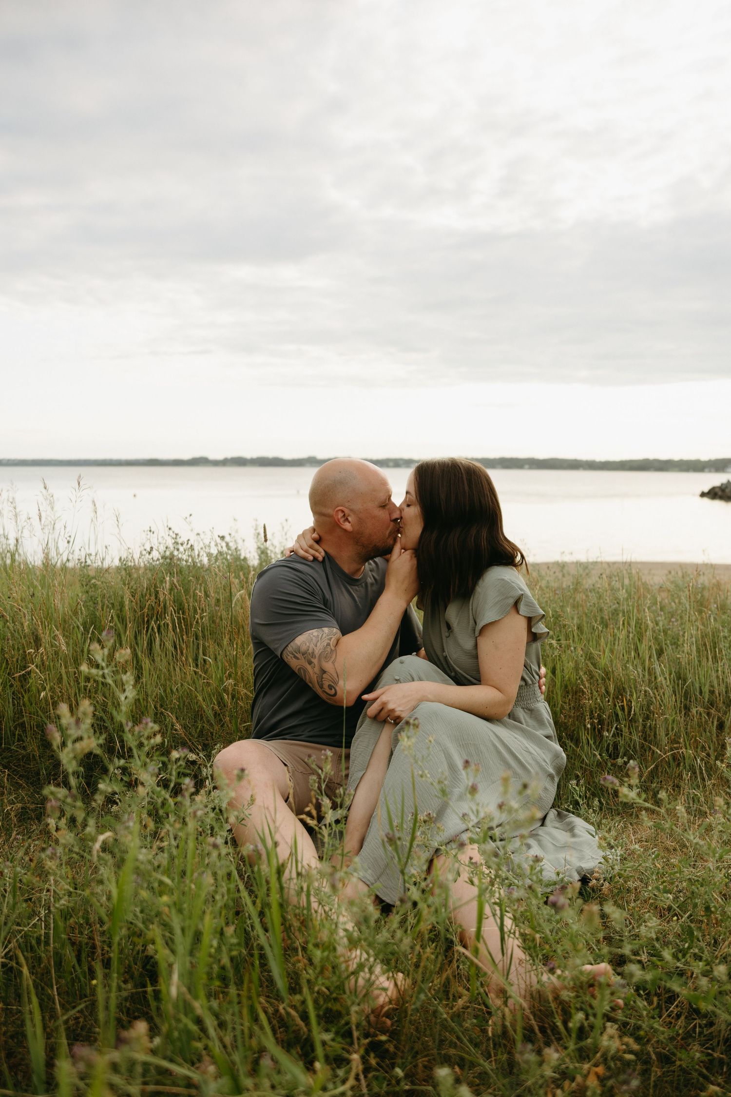A couple shares an intimate moment while sitting together in a field of tall grass near a lake on an overcast day.