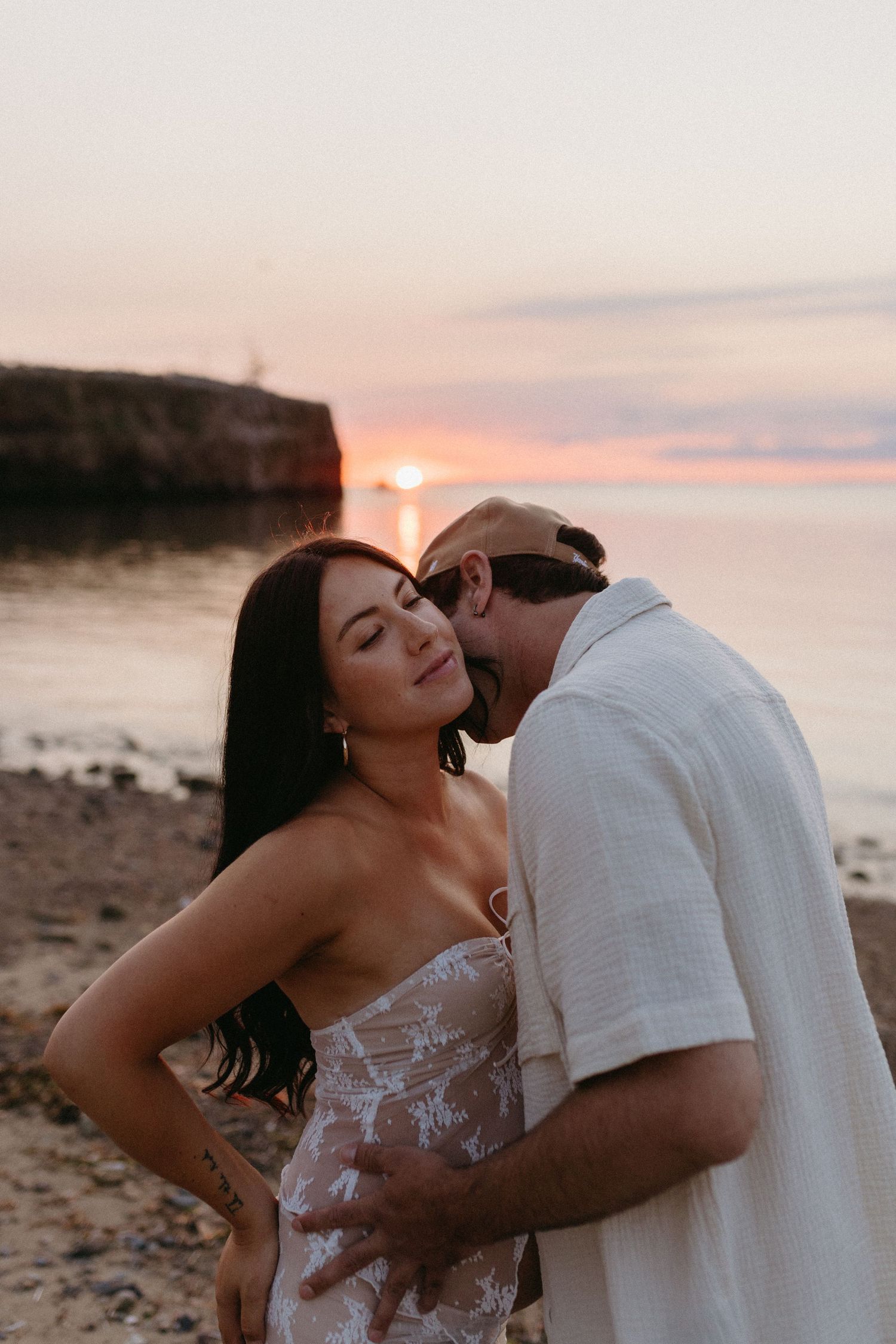 A romantic couple shares an intimate moment on a sandy beach during golden hour with cliffs in the distance.