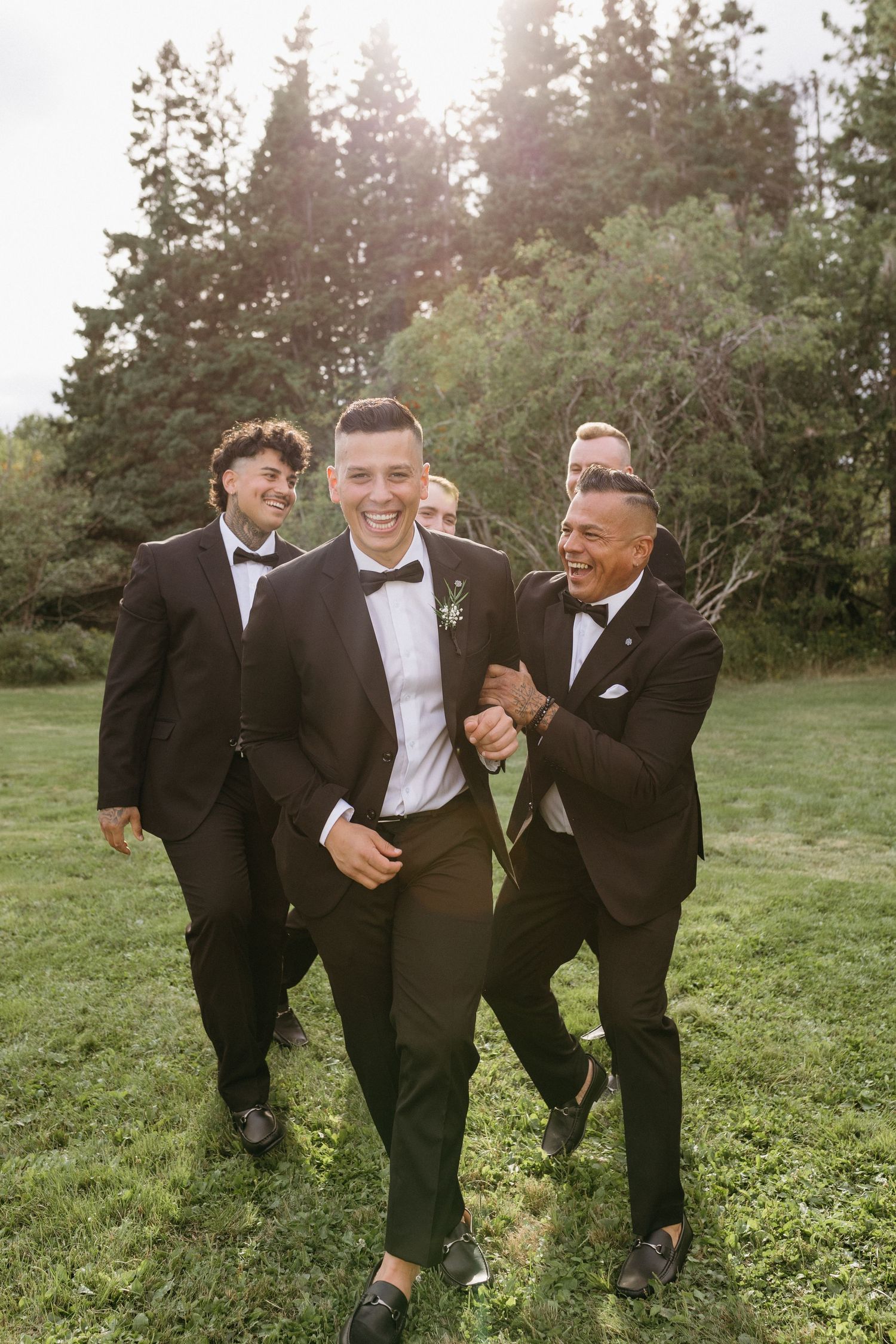 Groomsmen in black tuxedos share candid laughing moments in a grassy field during sunset.