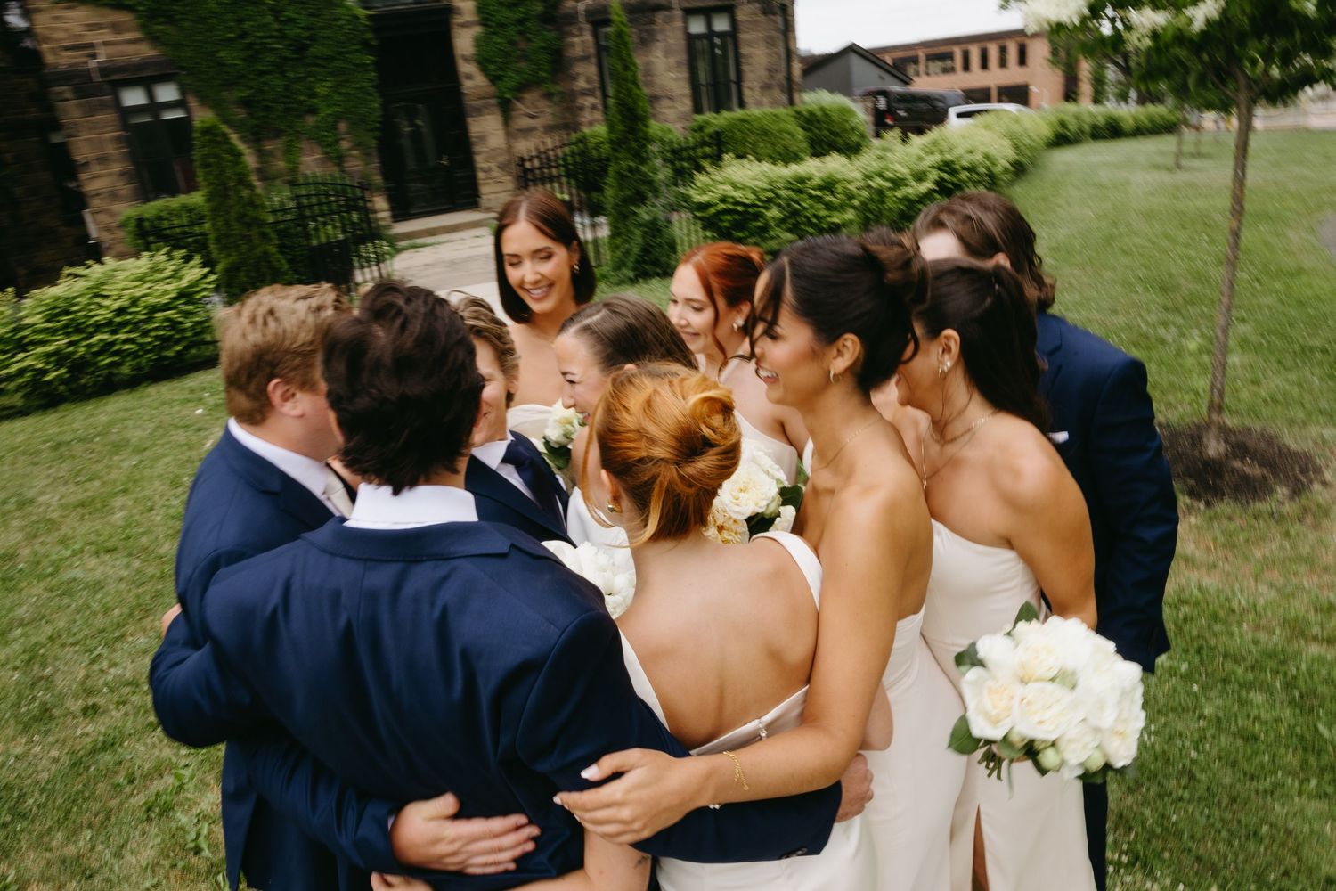 Wedding party gathers in a close circle on a grassy lawn outside a brick building, holding white bouquets.