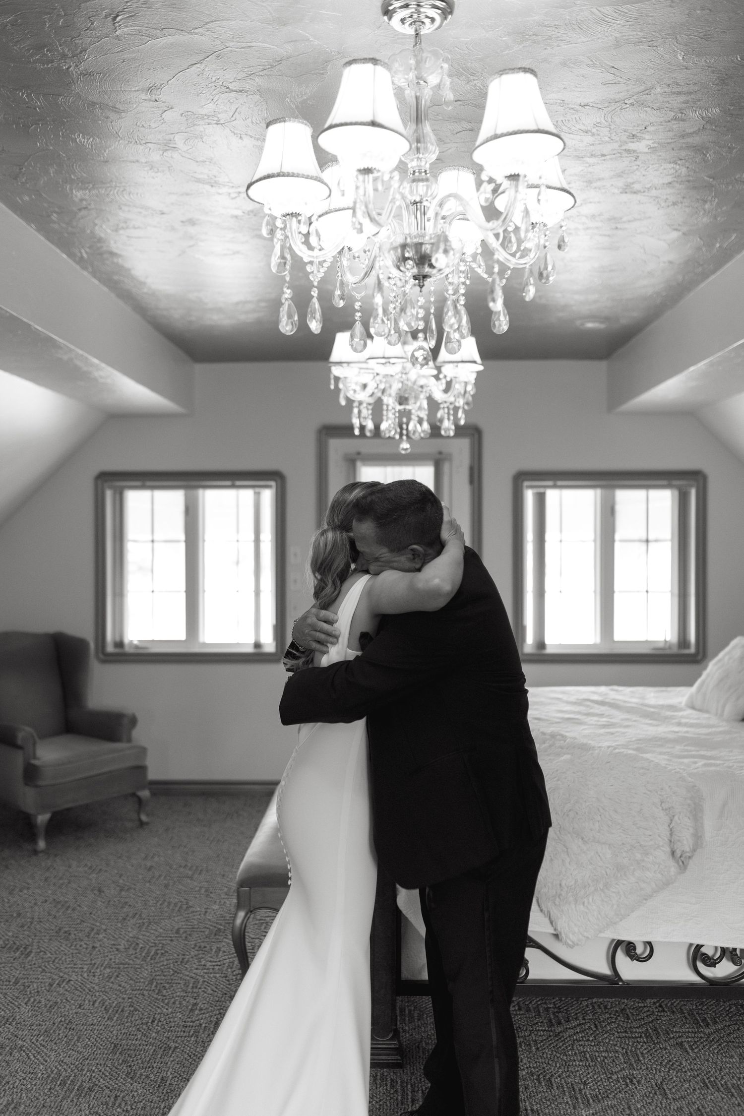 A romantic embrace under an elegant crystal chandelier in a bright room with windows and white decor.