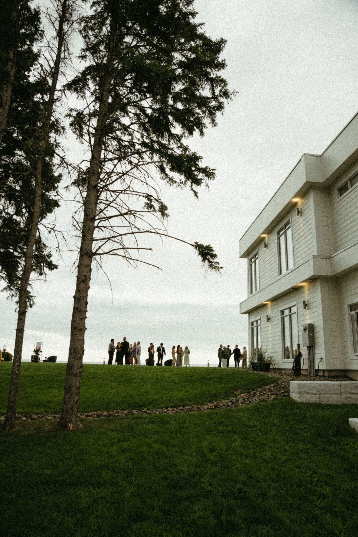 A white modern house on a grassy hill with tall pine trees and silhouetted figures gathered on the horizon.