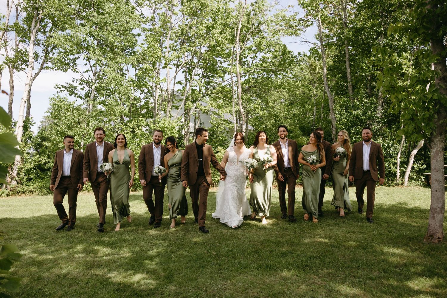 Wedding party poses under tall trees in a lush green outdoor setting during summer celebration.