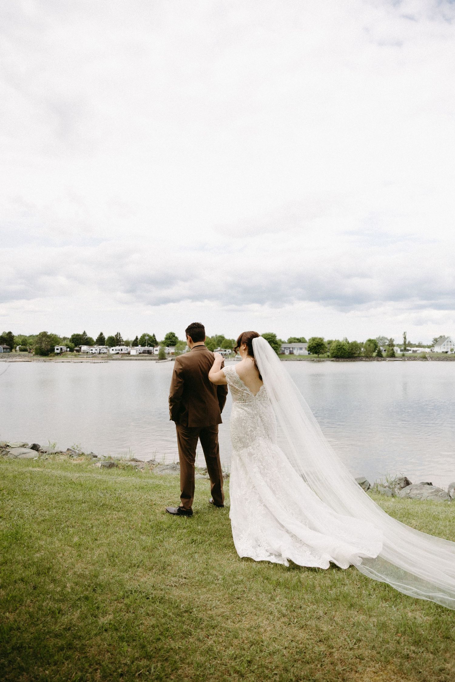 A long white wedding veil trails behind a couple on grassy waterfront grounds as storm clouds gather overhead.