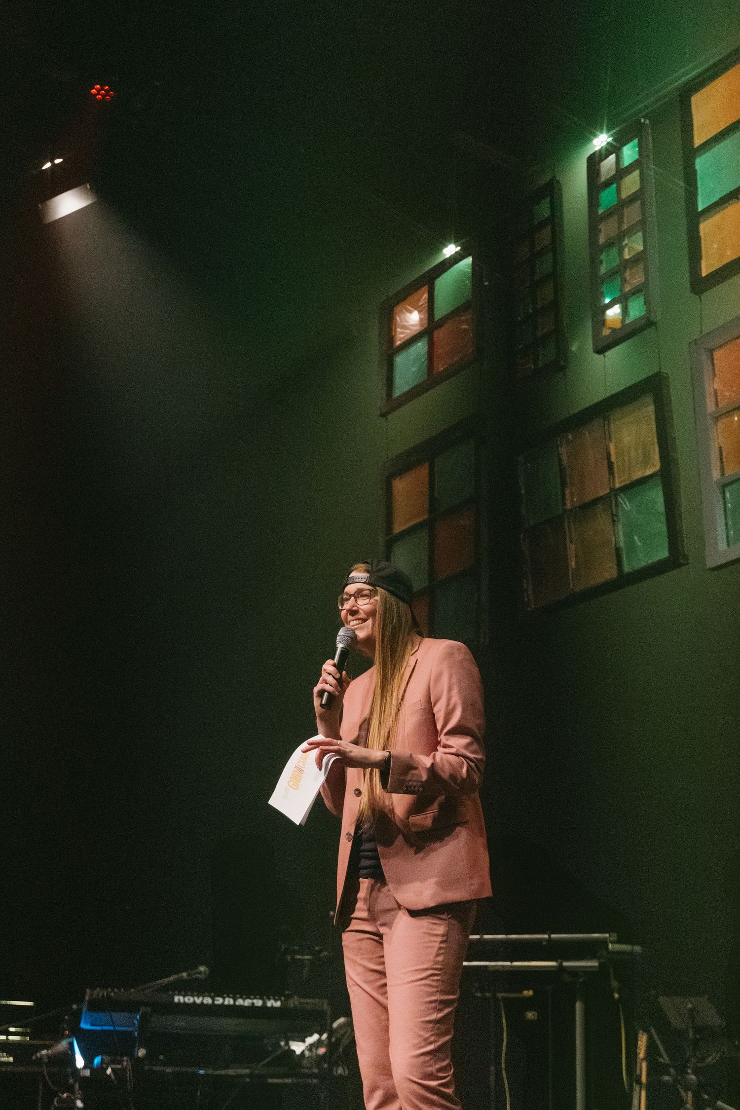A performer in a pink suit stands on stage under moody green lighting during a live performance.