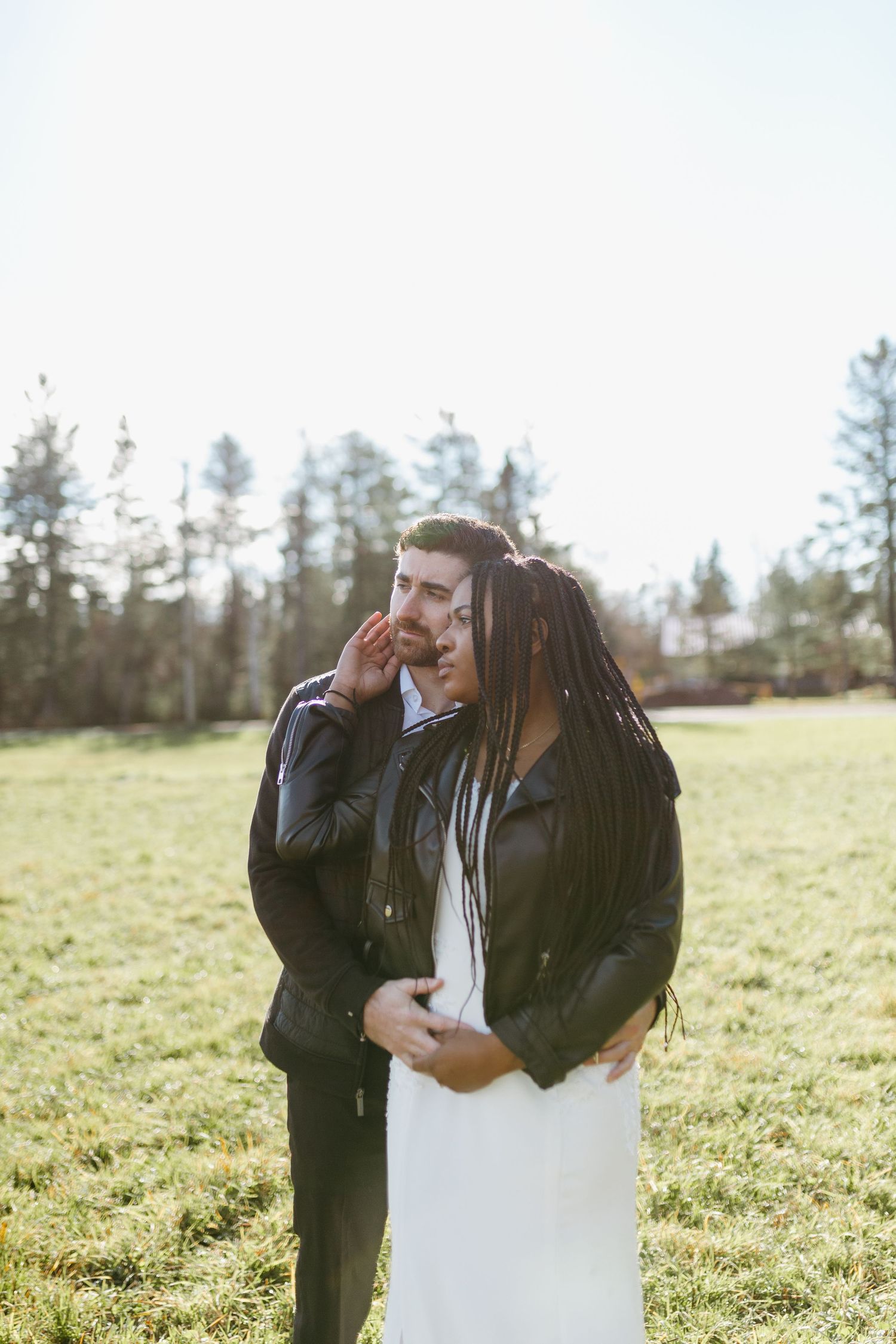 A couple shares a tender moment together in a sunny meadow surrounded by evergreen trees.
