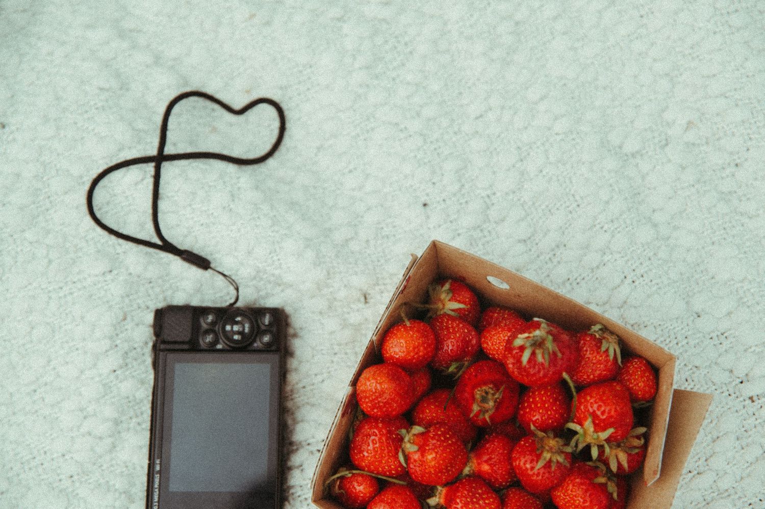 Fresh red strawberries spill out of a brown paper bag next to a vintage camera on a light green surface.