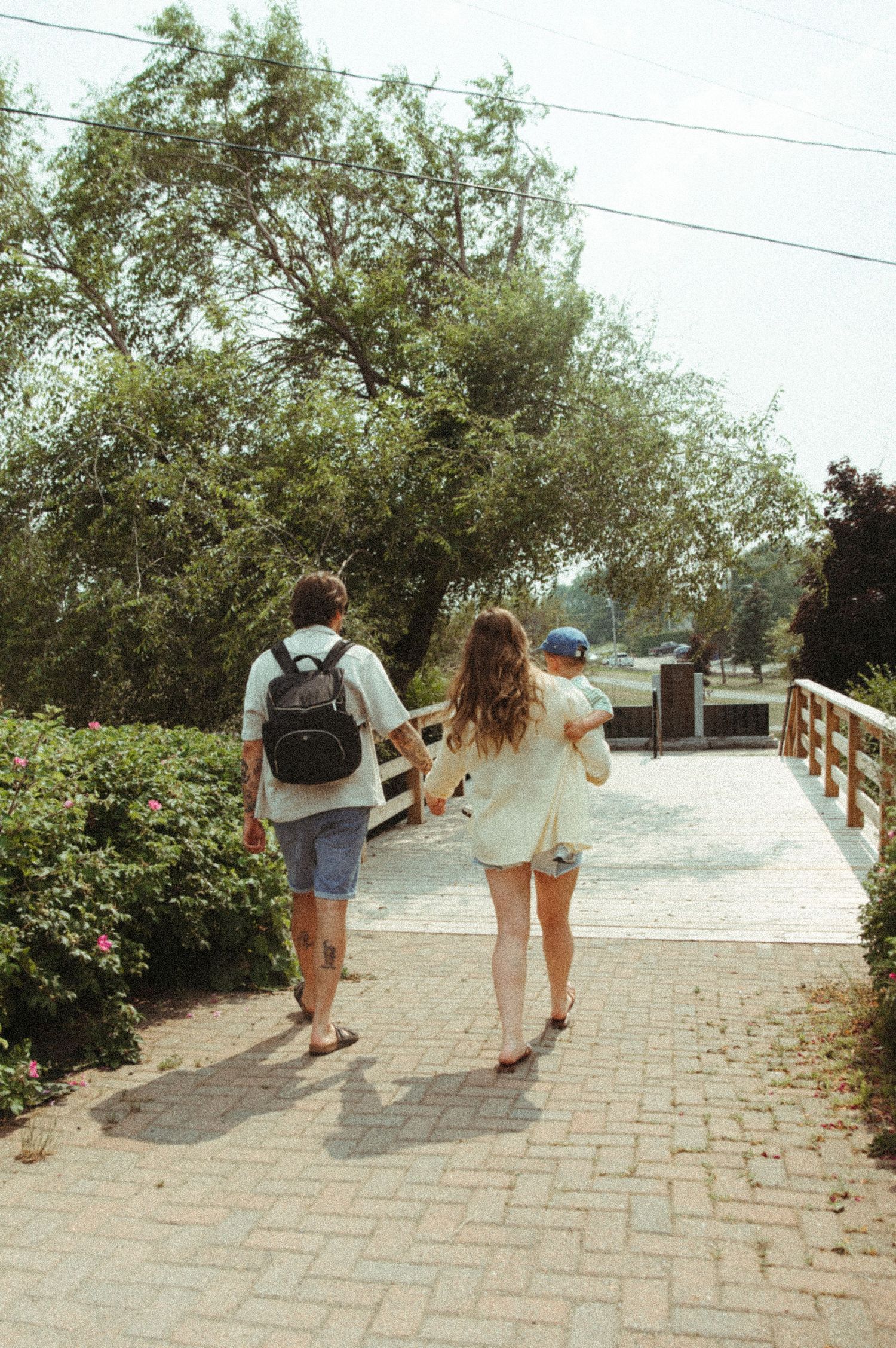 Two people walking along a brick paved pathway surrounded by greenery and trees on a sunny day.
