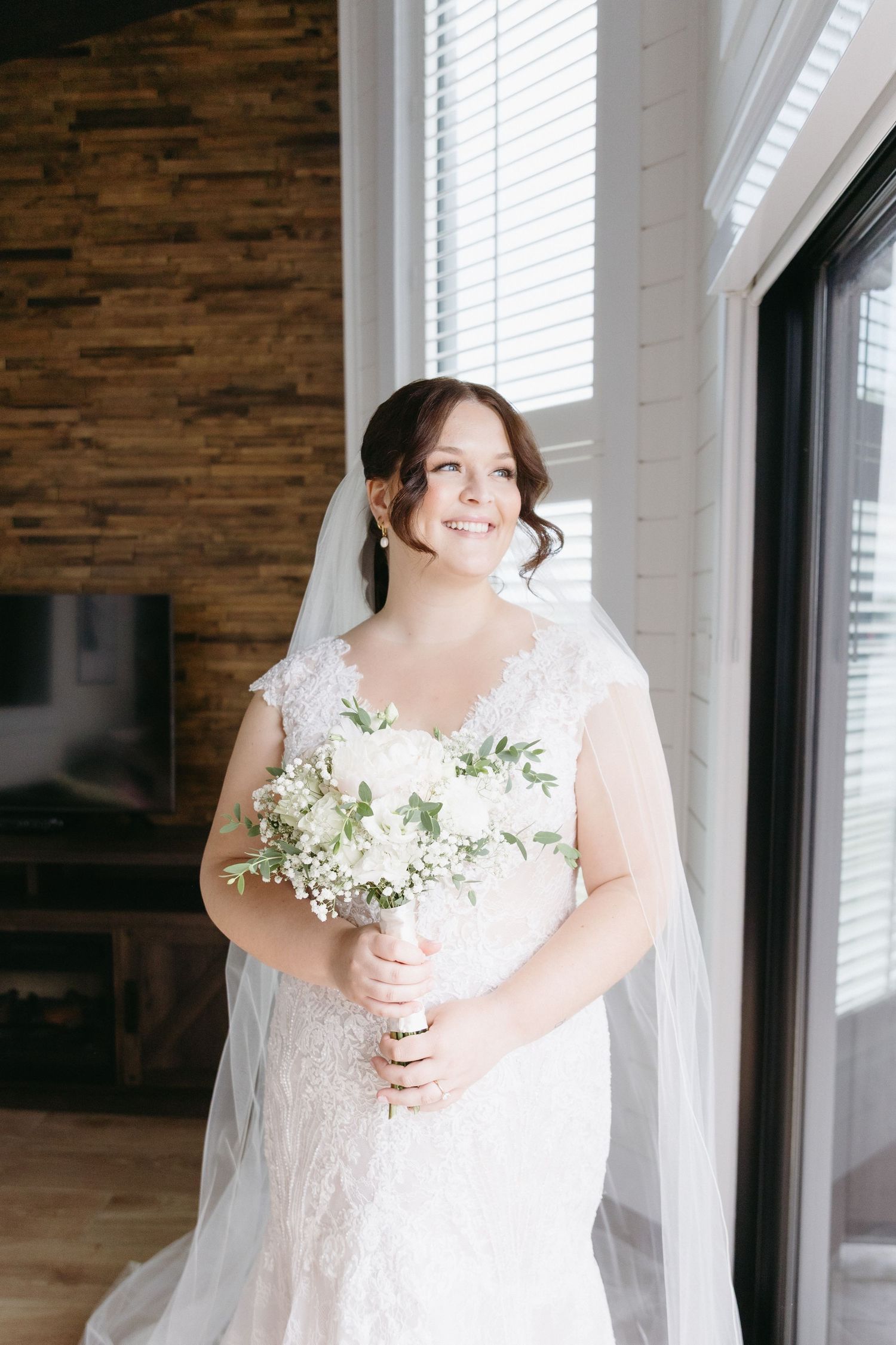 A bride in a white wedding dress holds a delicate bouquet of white flowers against a rustic wooden wall.