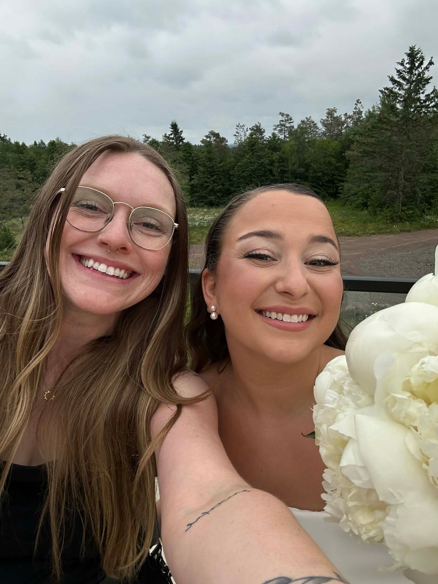 Two friends taking a selfie outdoors with evergreen trees and white peonies visible in the background.