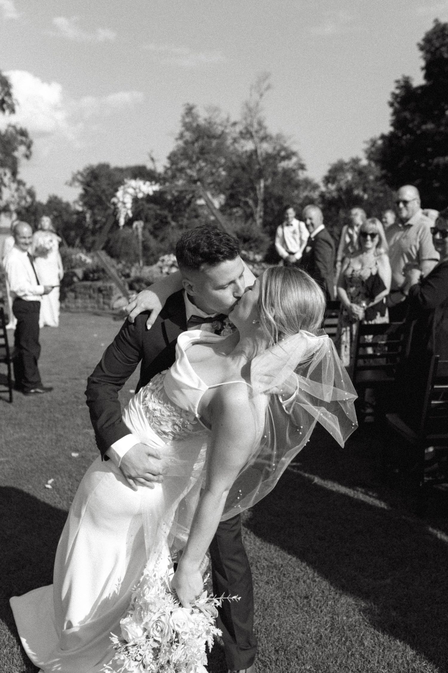 Black and white photo of wedding guests gathered outdoors for ceremony seating on lawn.