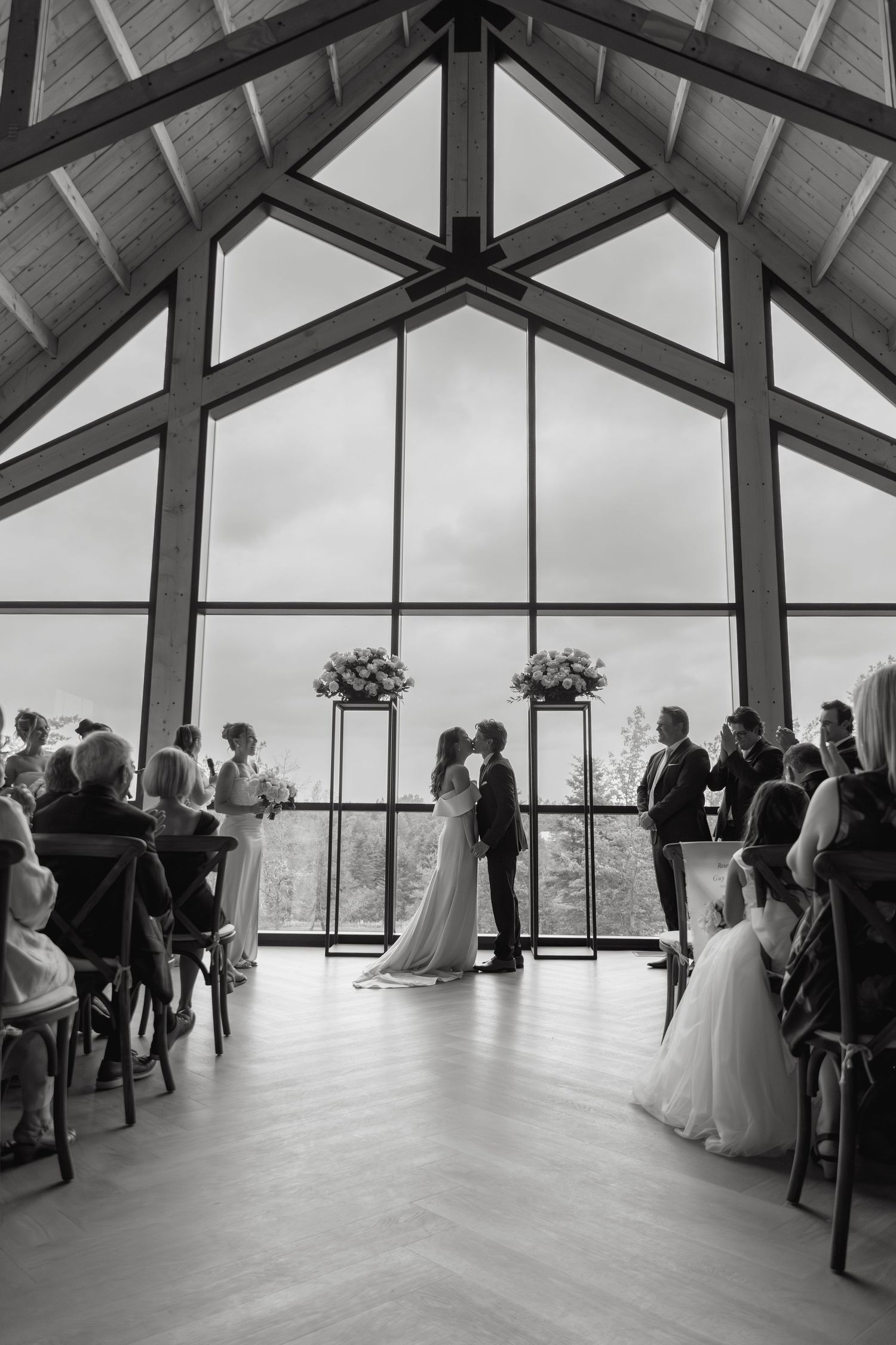 Wedding ceremony taking place in modern glass-walled venue with panoramic views and guests gathered at the windows.