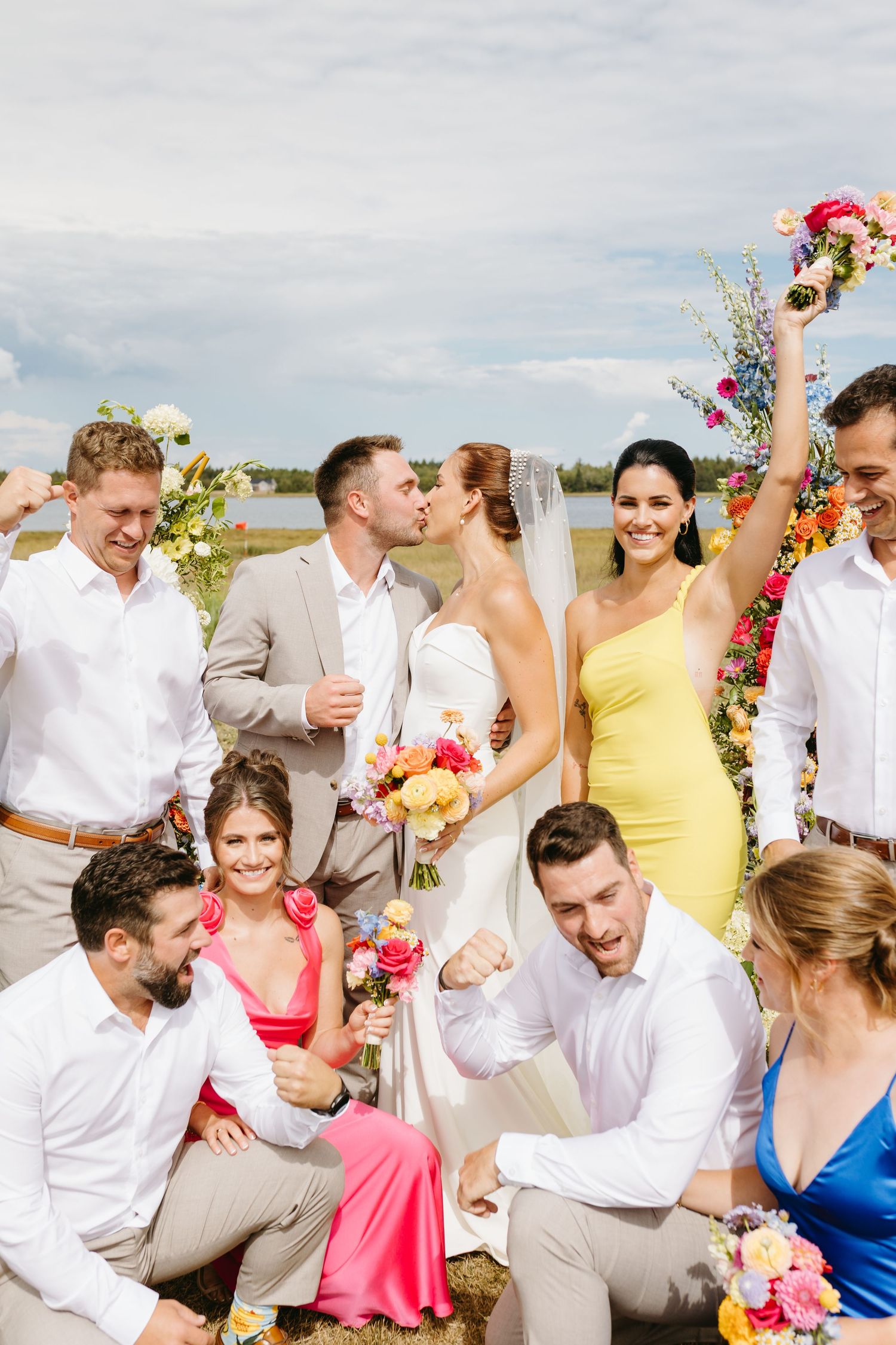 Wedding party in bright summer attire celebrates on beach with colorful floral bouquets and joyful expressions.