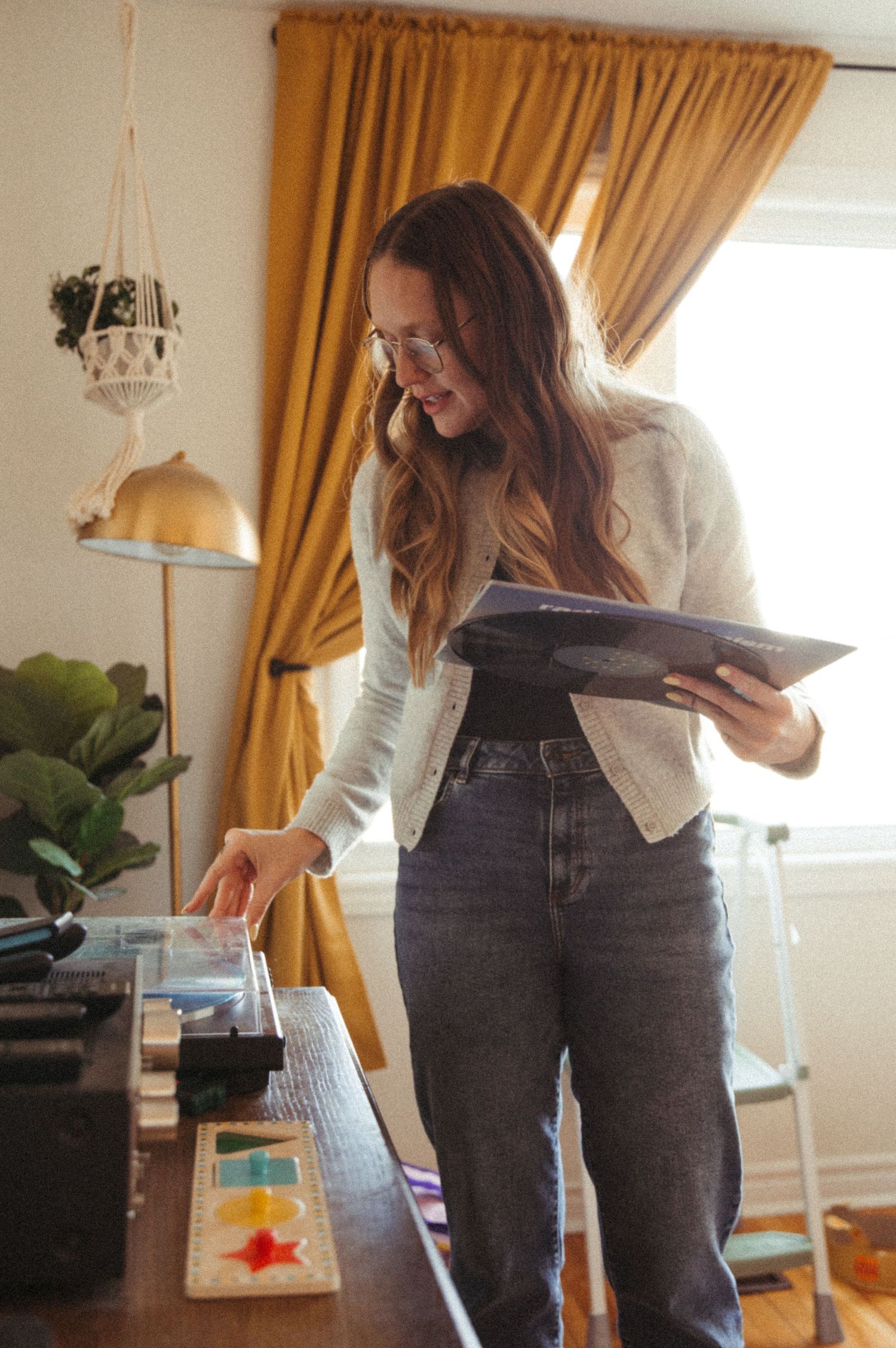 A person in a white cardigan and jeans looks at papers while standing in a warmly lit room with gold curtains and plants.