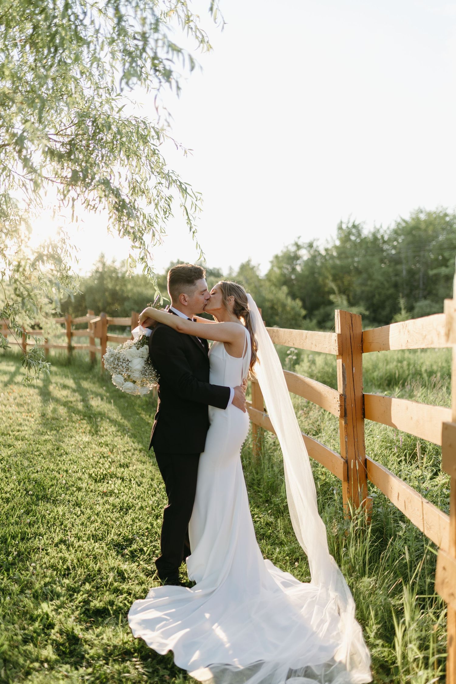 A romantic wedding couple embraces near a wooden fence in a sunlit meadow with a flowing white dress and veil.