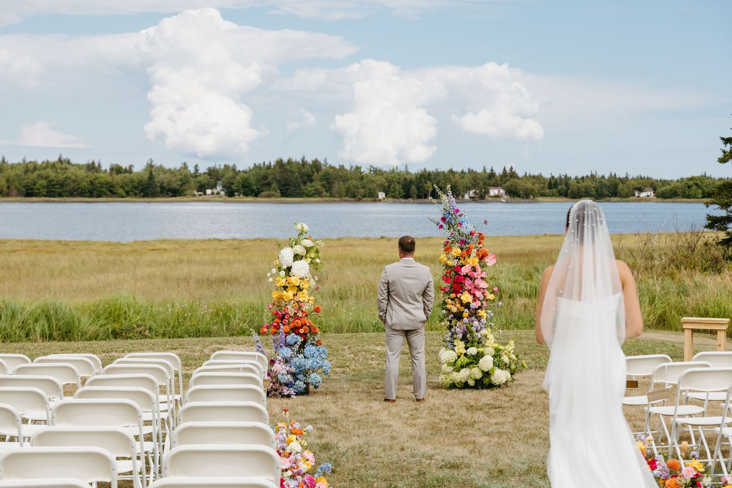Outdoor lakeside wedding ceremony setup with white chairs and colorful floral arrangements beside a serene waterfront.