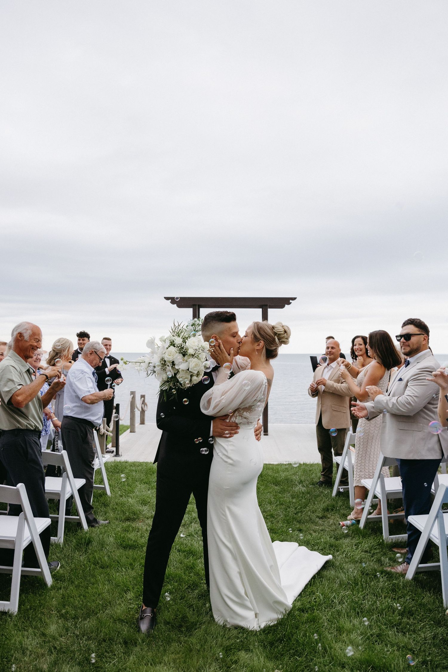 Wedding guests dance in pairs on a grassy lawn with white chairs and a cloudy sky backdrop during an outdoor celebration.