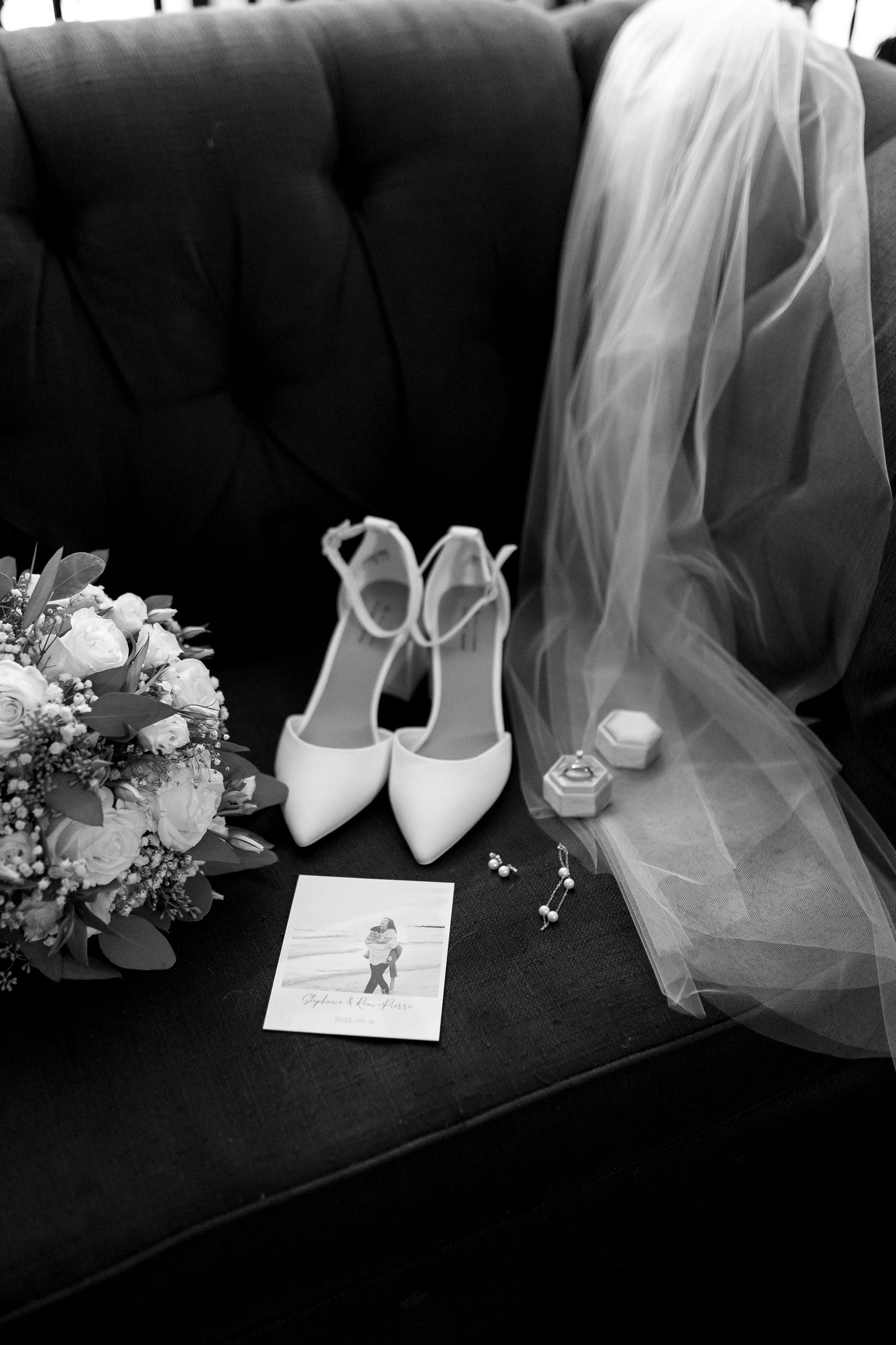 Wedding details arranged on dark couch including white shoes, veil, bouquet and a small photo.